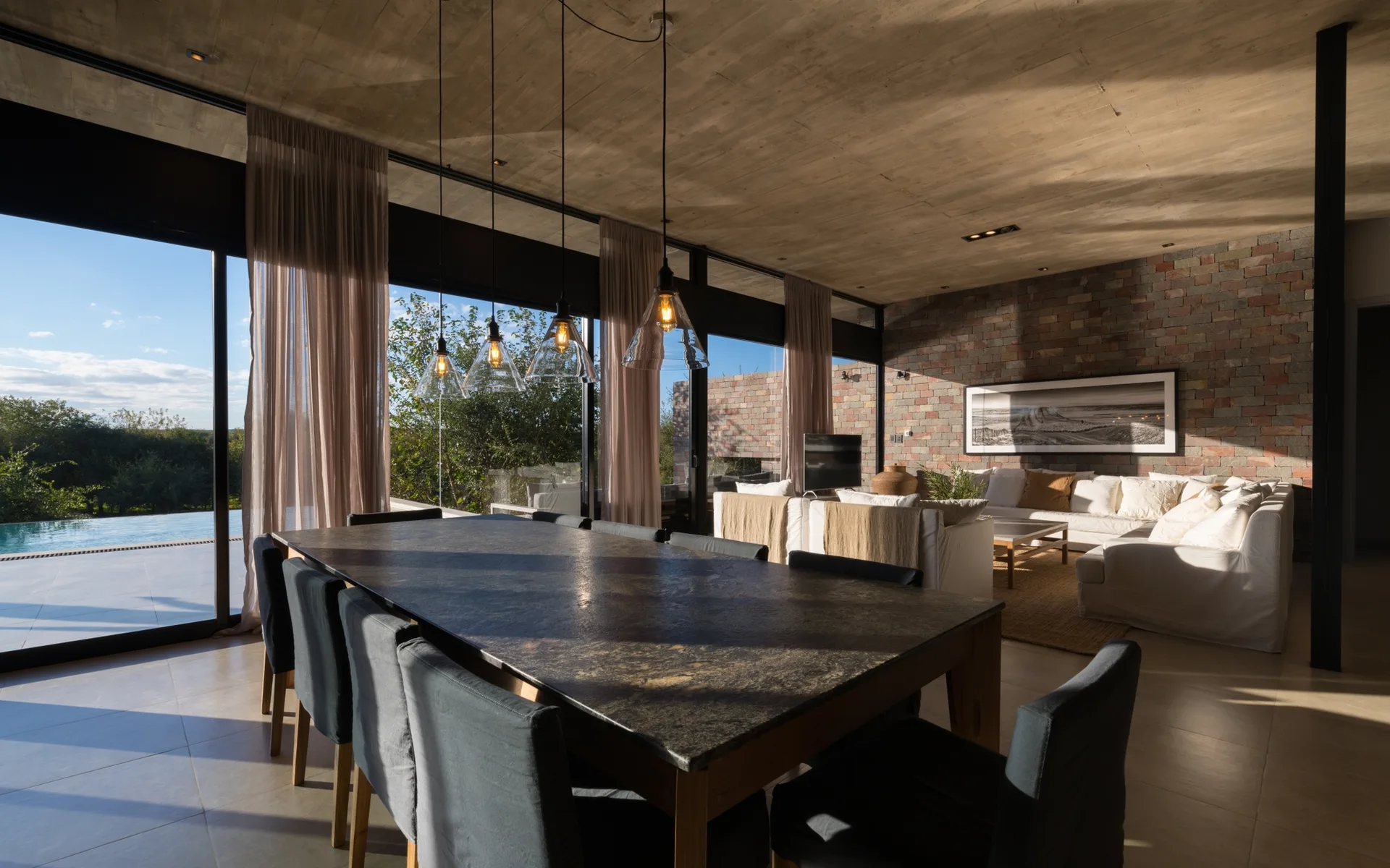 An open-plan interior features a dark stone dining table with grey chairs beneath a raw concrete ceiling and adjacent to a living area with a white sofa.