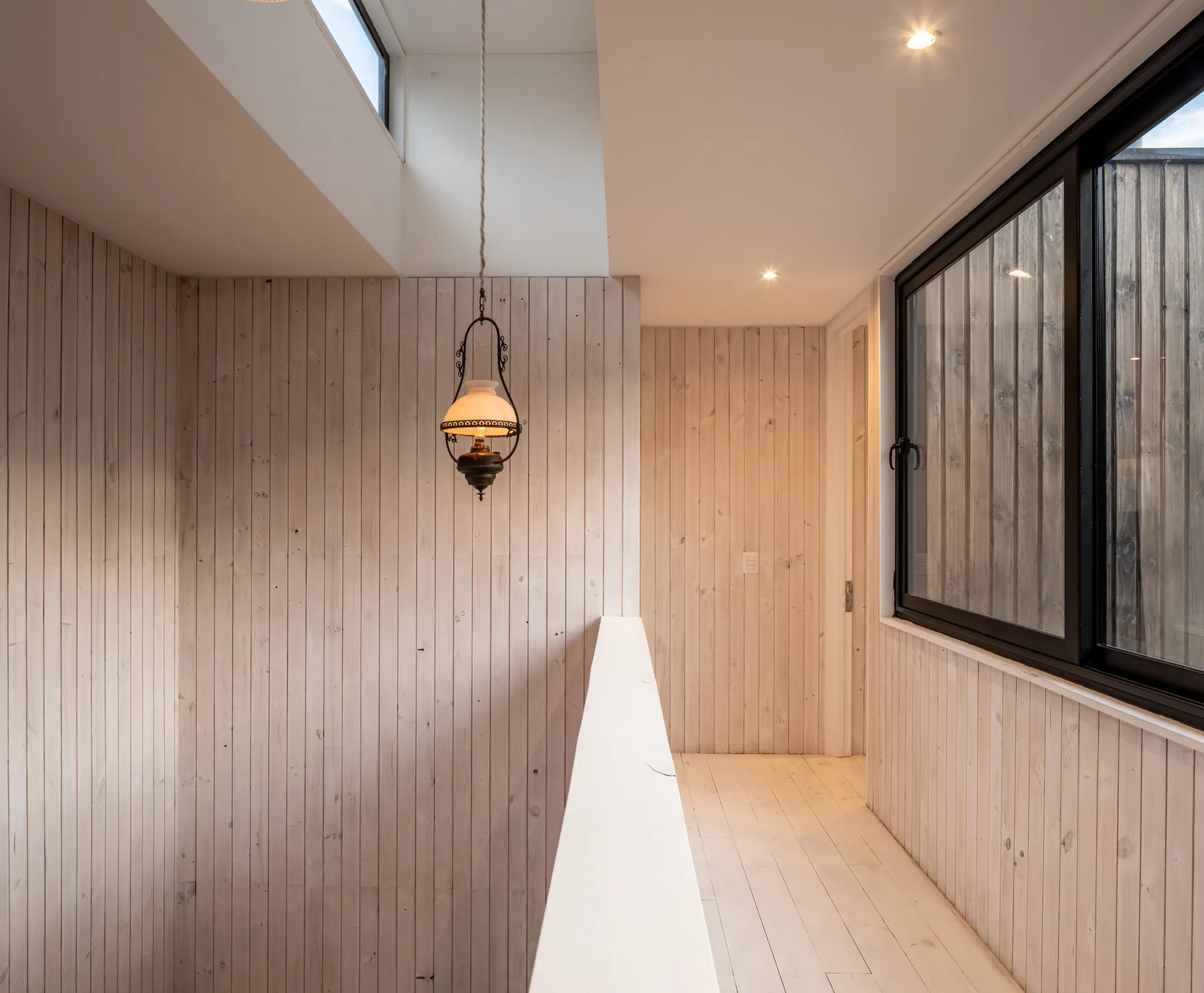 A minimalist landing featuring whitewashed vertical wood paneled walls, a matching wood floor, a skylight, and a hanging vintage pendant light.