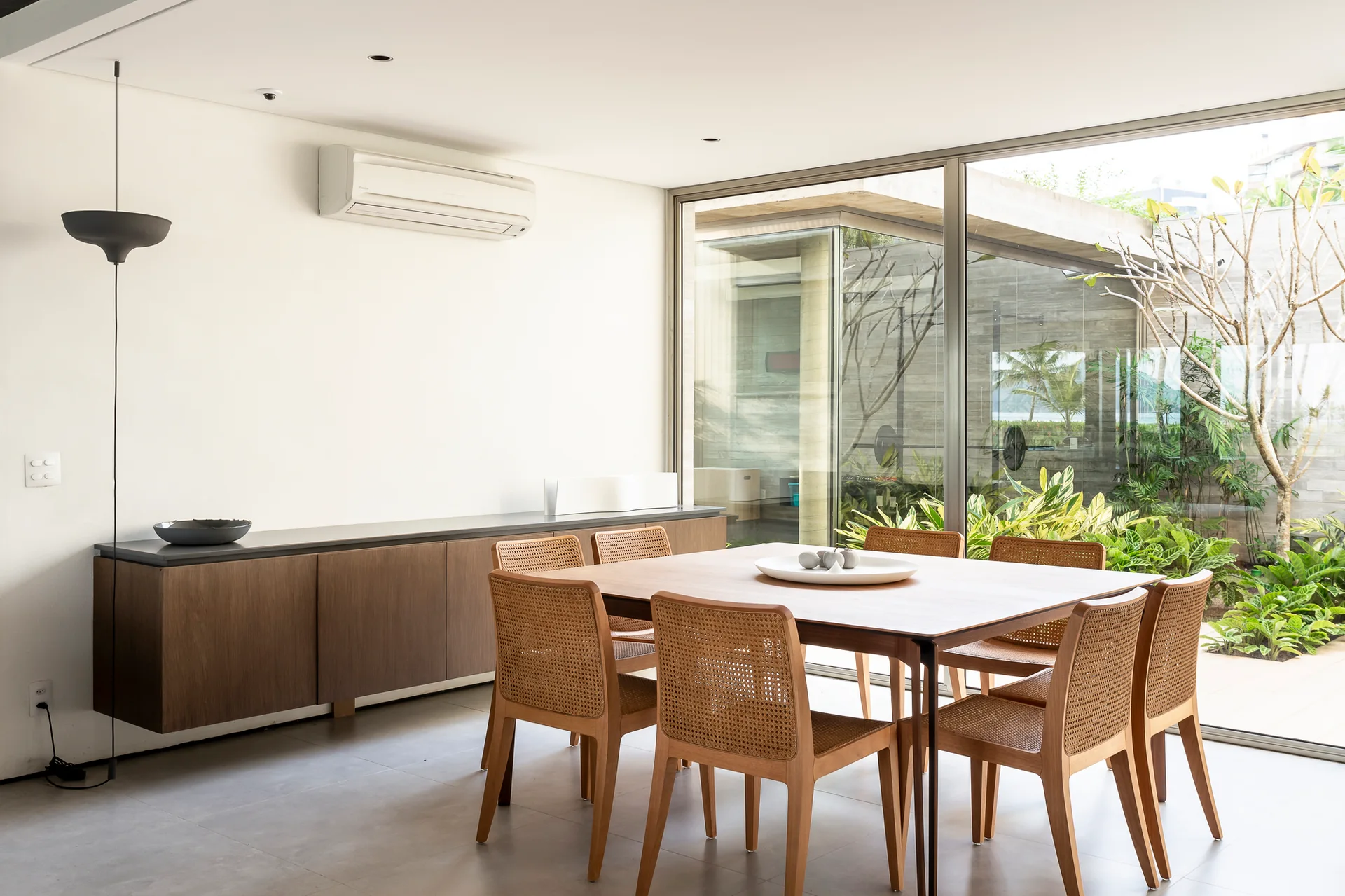 A minimalist dining room features a wooden table, woven rattan chairs, a wall-mounted floating cabinet, and sliding glass doors opening to a garden.