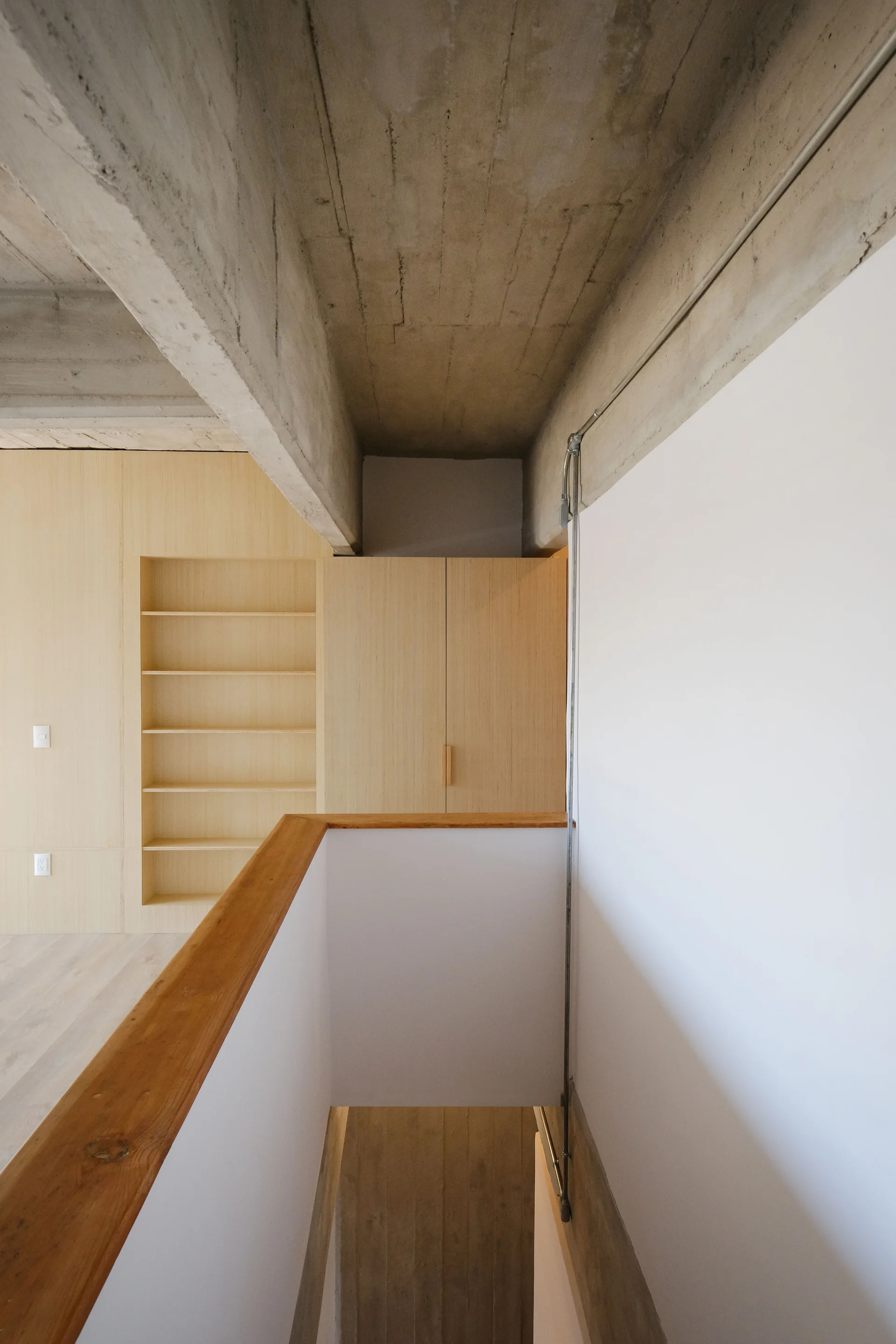 A view looking down a minimalist staircase with a wooden handrail, featuring light wood built-in cabinetry and an exposed concrete ceiling.