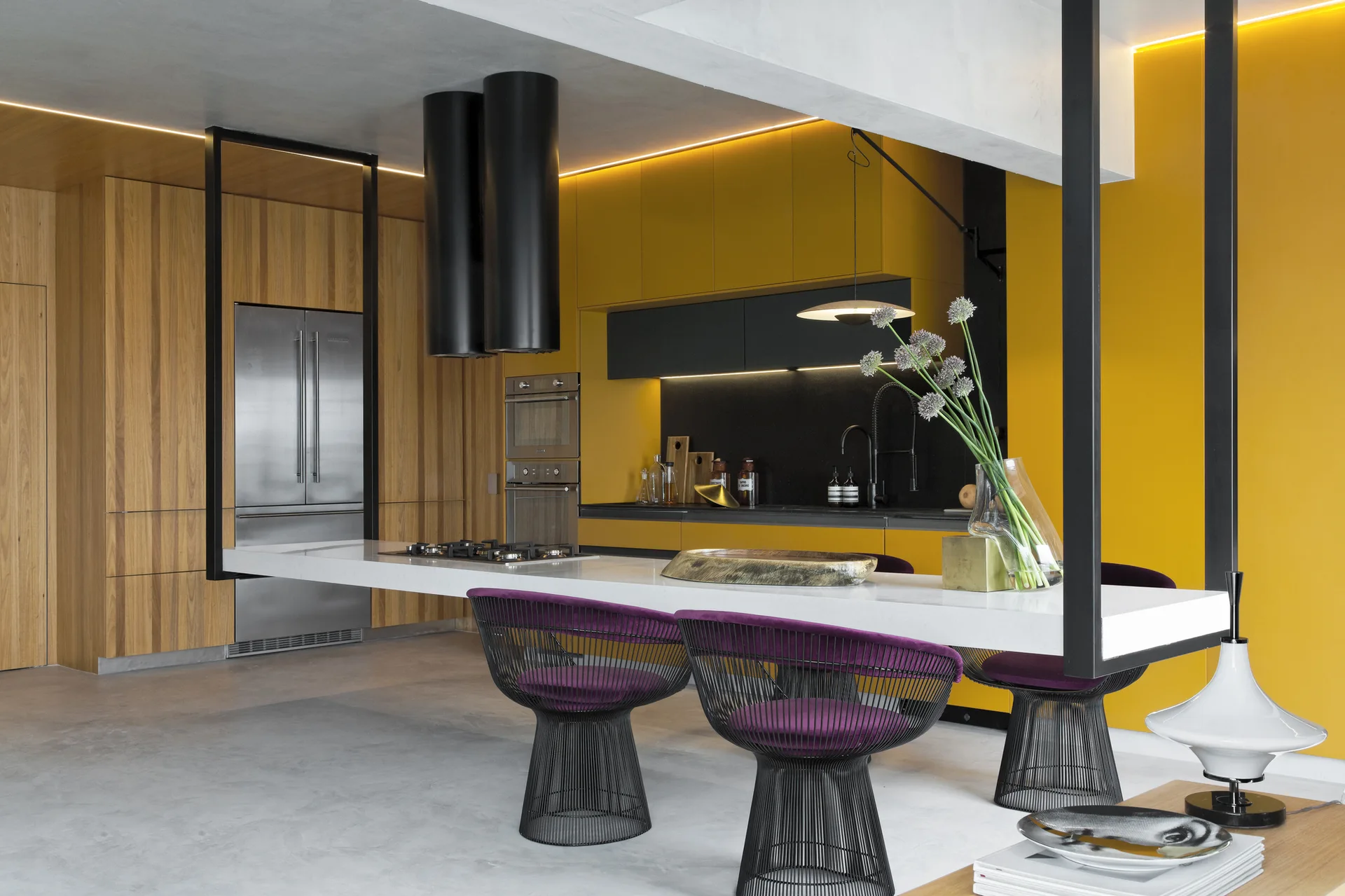 A modern kitchen featuring a bright yellow accent wall, wood cabinetry, a long white cantilevered island, and purple wire chairs.