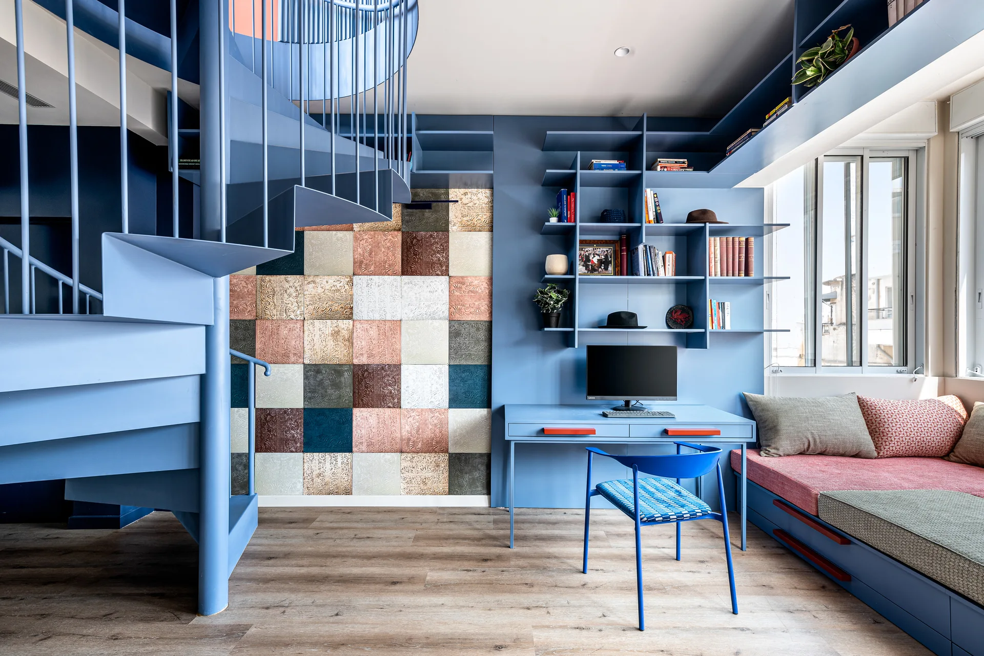 A blue home office featuring a matching blue metal spiral staircase, integrated desk and shelving, and a built-in daybed over light wood flooring.