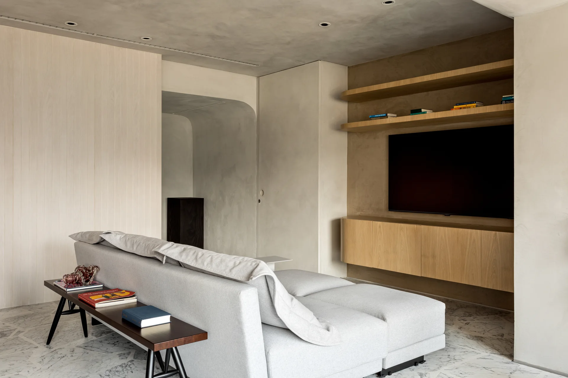 A minimalist living room features a grey modular sofa, a dark wooden bench, veined marble flooring, and built-in light wood media cabinetry.