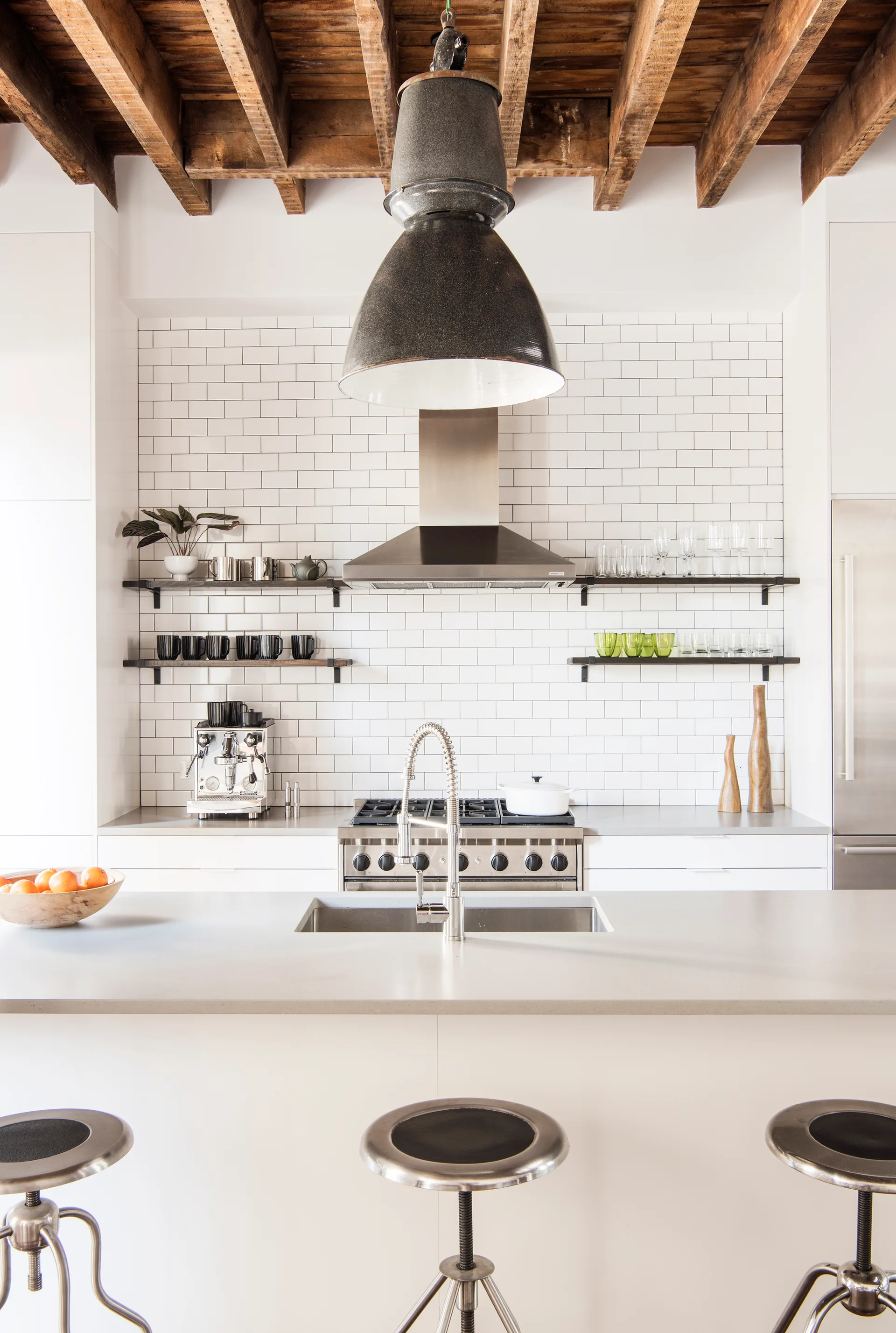 An industrial kitchen with a white island, metal stools, a subway tile backsplash, and a large pendant hanging from exposed wooden ceiling beams.