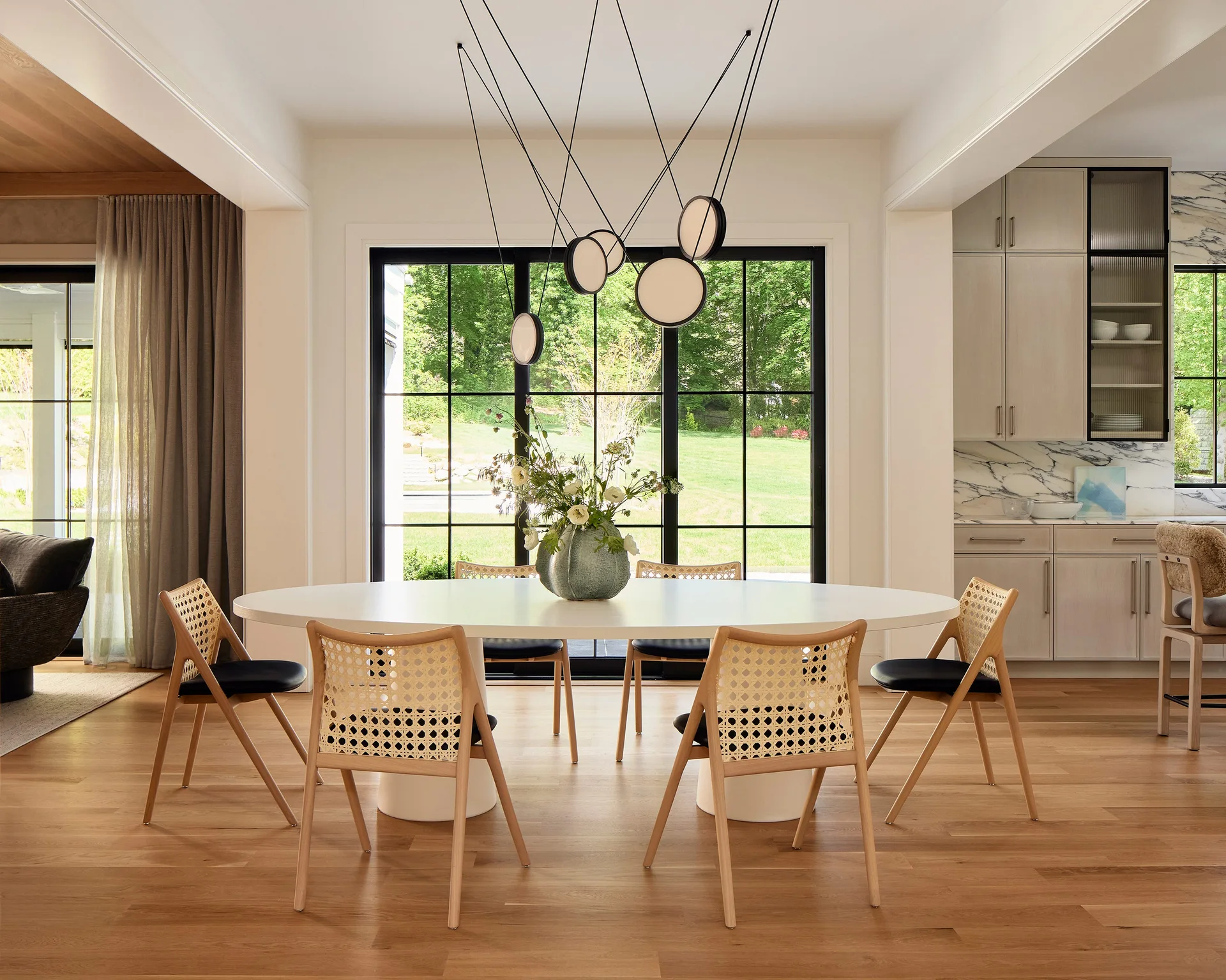 An oval white dining table with light wood cane chairs is positioned in front of large black-framed glass doors leading outside.