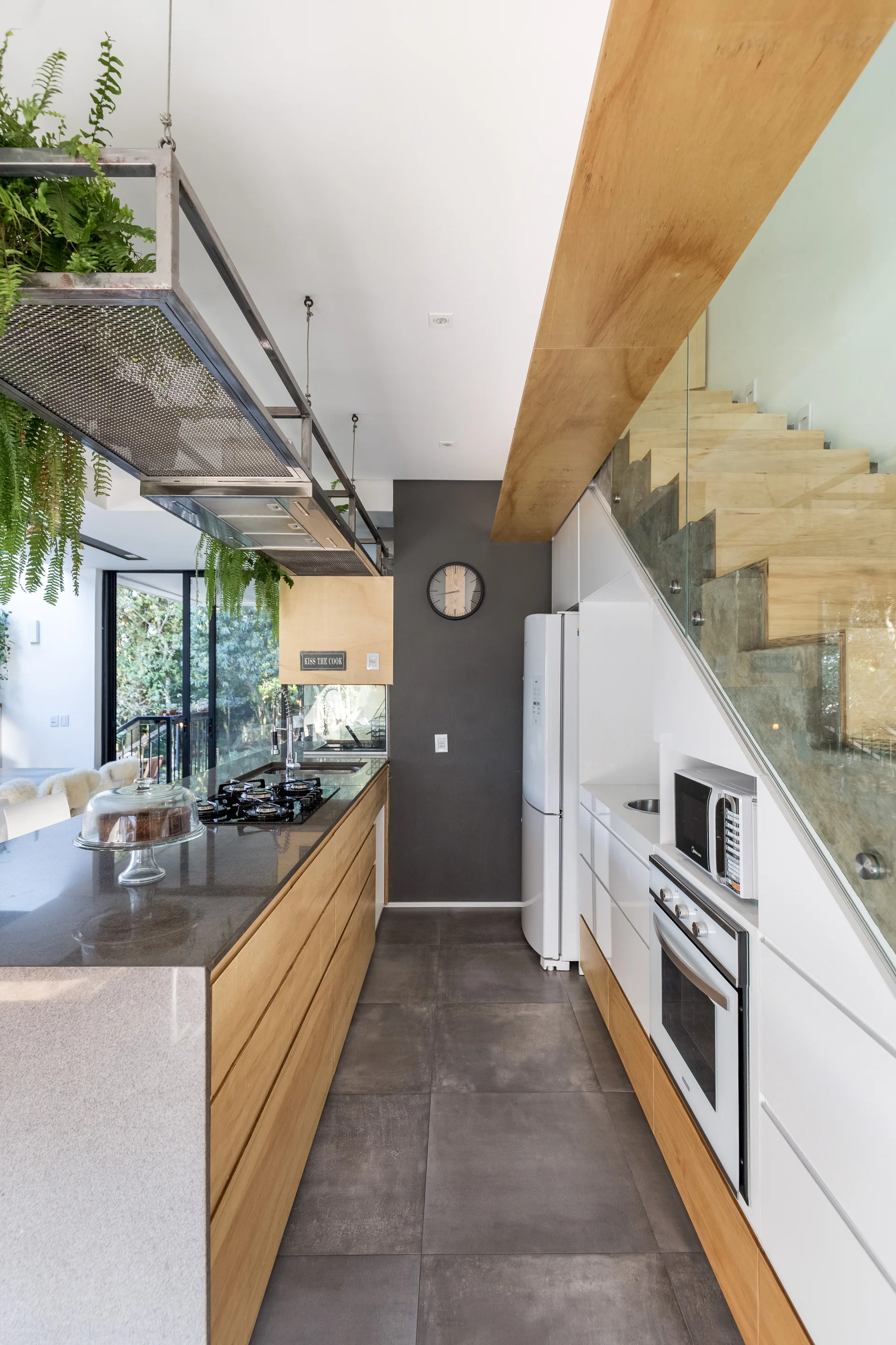A modern galley kitchen features a wood-paneled island, integrated under-stair appliances, and a hanging metal range hood with lush green ferns.