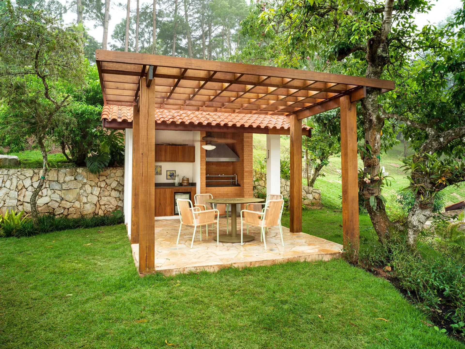 A covered outdoor kitchen featuring a wooden pergola, brick barbecue, stone patio floor, and a small dining table with four chairs set on a green lawn.