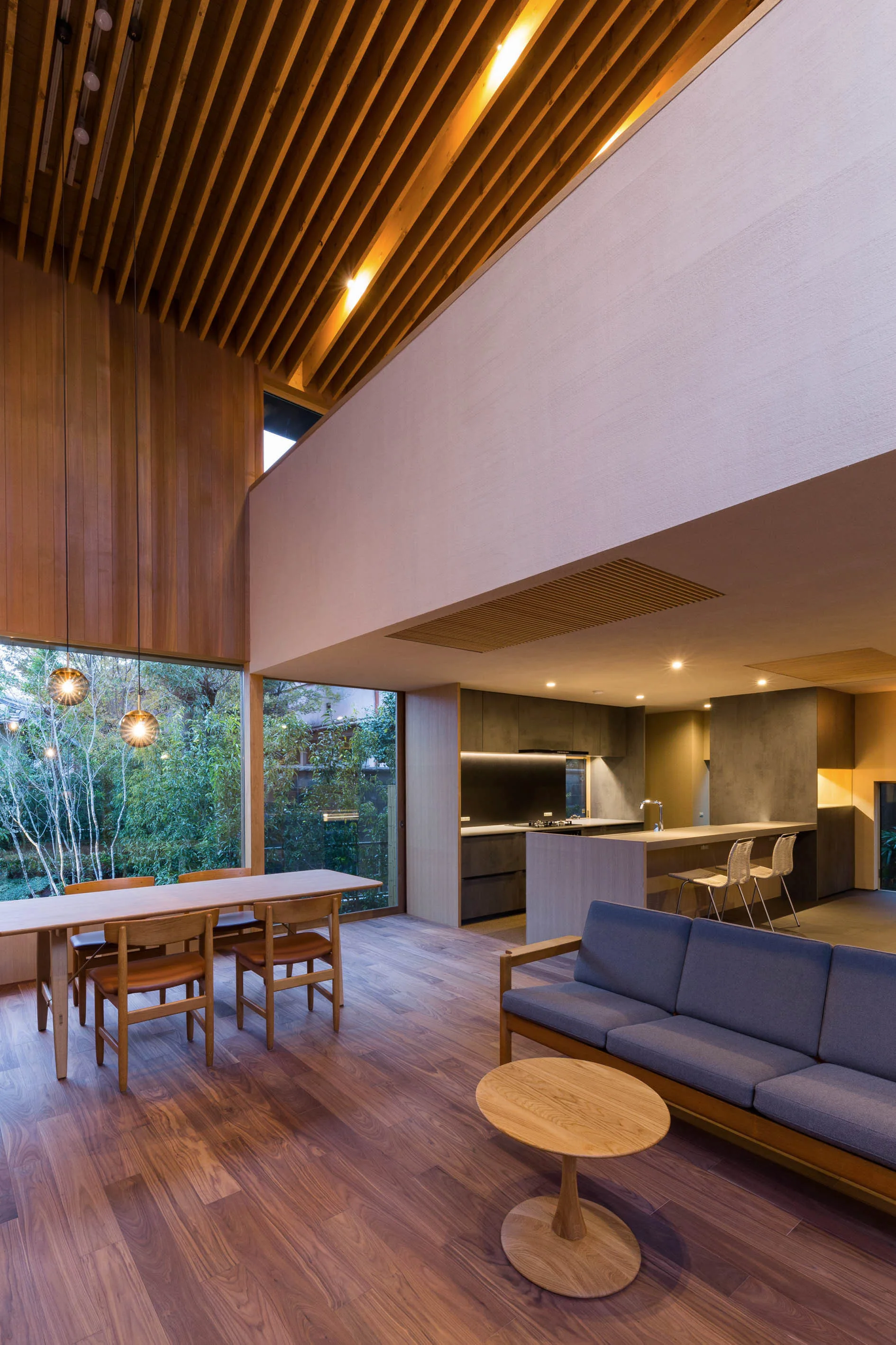 An open-concept living space features a soaring timber slat ceiling, a wood dining table, a grey sofa, and a kitchen with dark cabinetry.