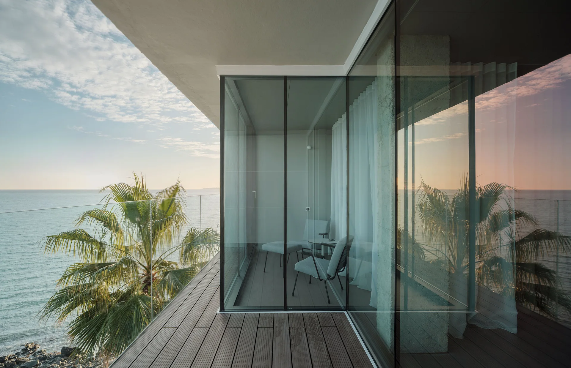 A modern oceanfront balcony features dark wood plank decking, a transparent glass railing, and floor-to-ceiling glass walls reflecting the sky.