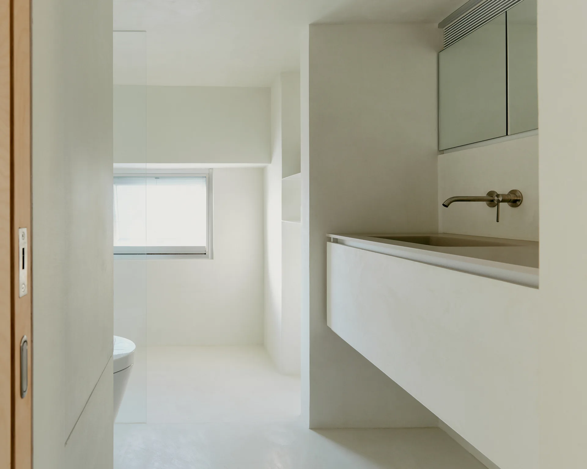 A minimalist white bathroom featuring a floating vanity with a wall-mounted faucet, recessed mirror, and seamless plaster finishes.