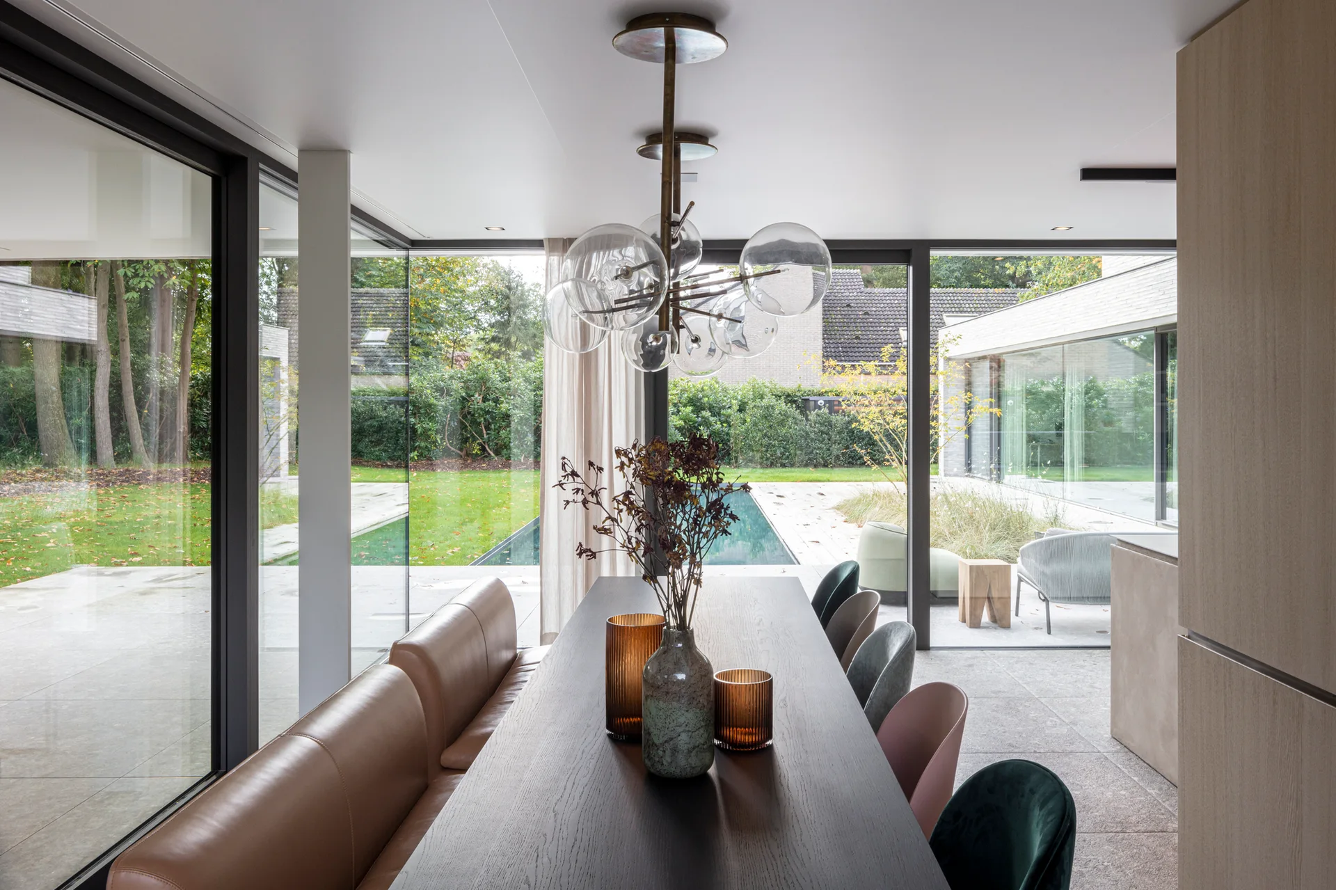 A modern dining room features a dark wood table with a tan leather bench and colorful chairs next to glass walls overlooking an outdoor pool.