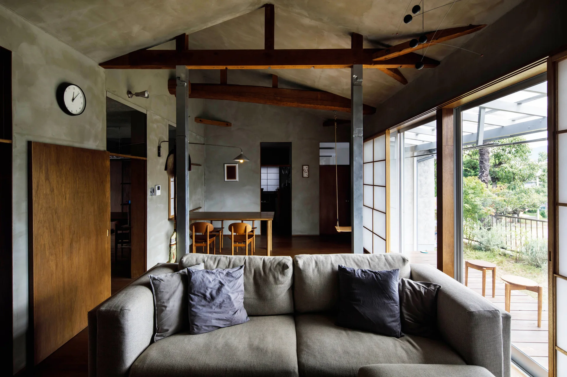 A minimalist living room features a grey sofa, exposed wood ceiling beams, matte concrete walls, and shoji-style sliding doors opening to a terrace.