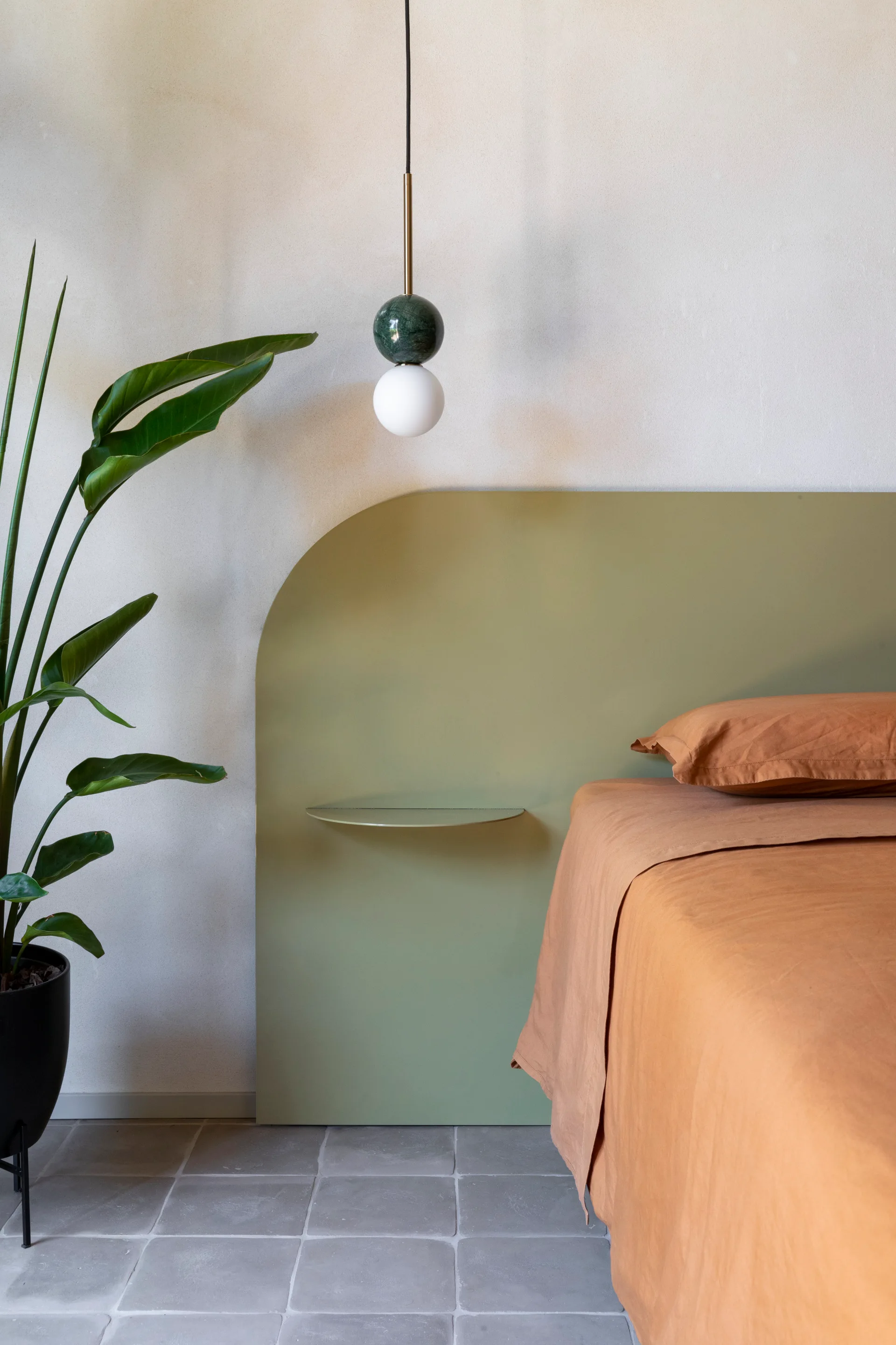 A minimalist bedroom featuring a curved sage green headboard with a built-in shelf, terracotta linens, and a marble sphere pendant light.