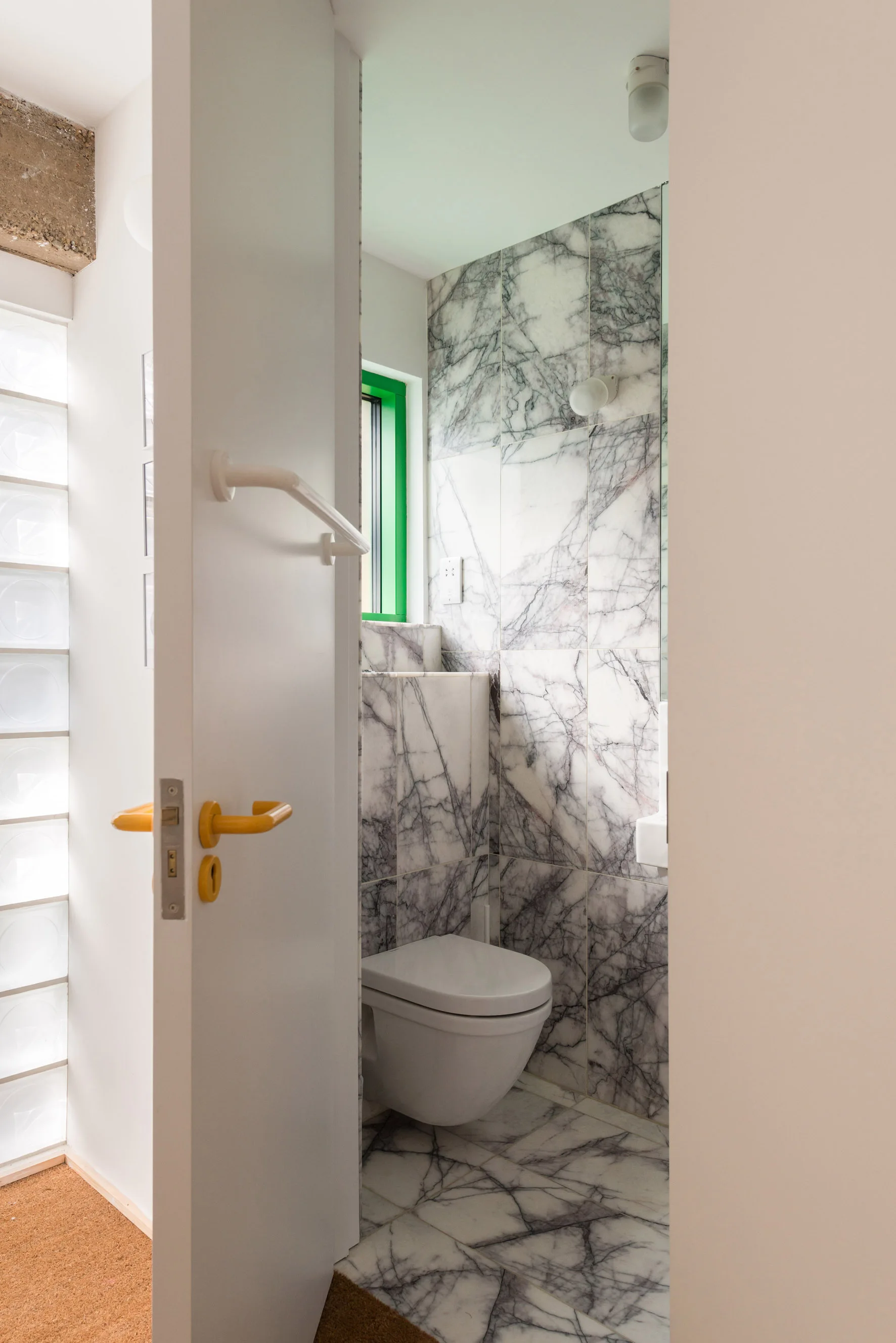 An open white door with a yellow handle reveals a small powder room clad in heavily veined marble tile with a bright green window frame.