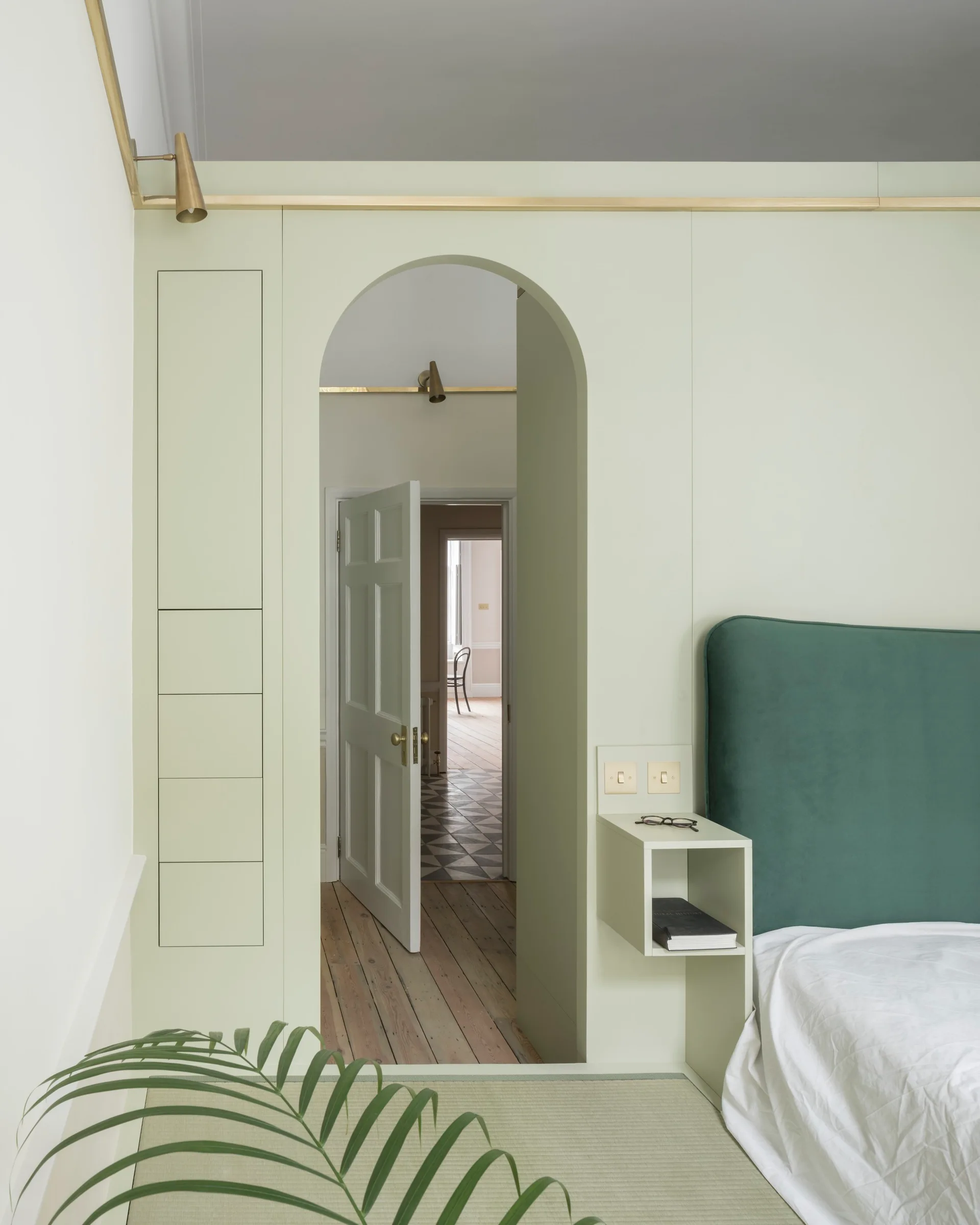 A bedroom features sage green custom built-in cabinetry with an arched doorway, an integrated floating shelf, and a deep green velvet headboard.