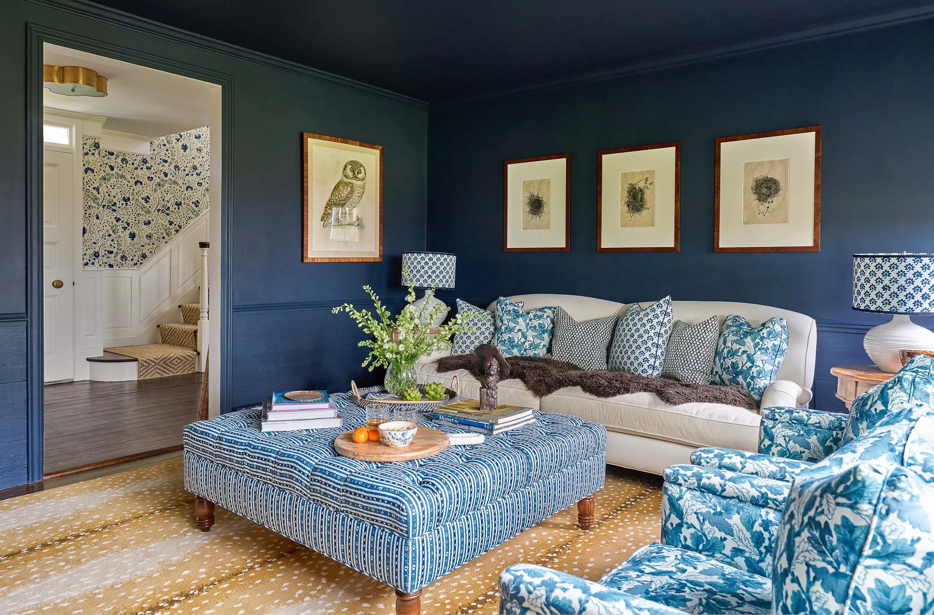 A navy blue living room features a white sofa with patterned pillows, a large tufted blue ottoman, and a fawn-patterned rug.