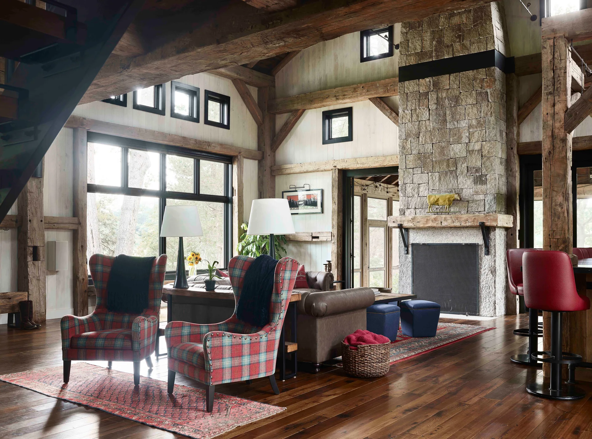 A rustic timber frame living room featuring a tall stone fireplace, plaid wingback chairs, and dark wood flooring.