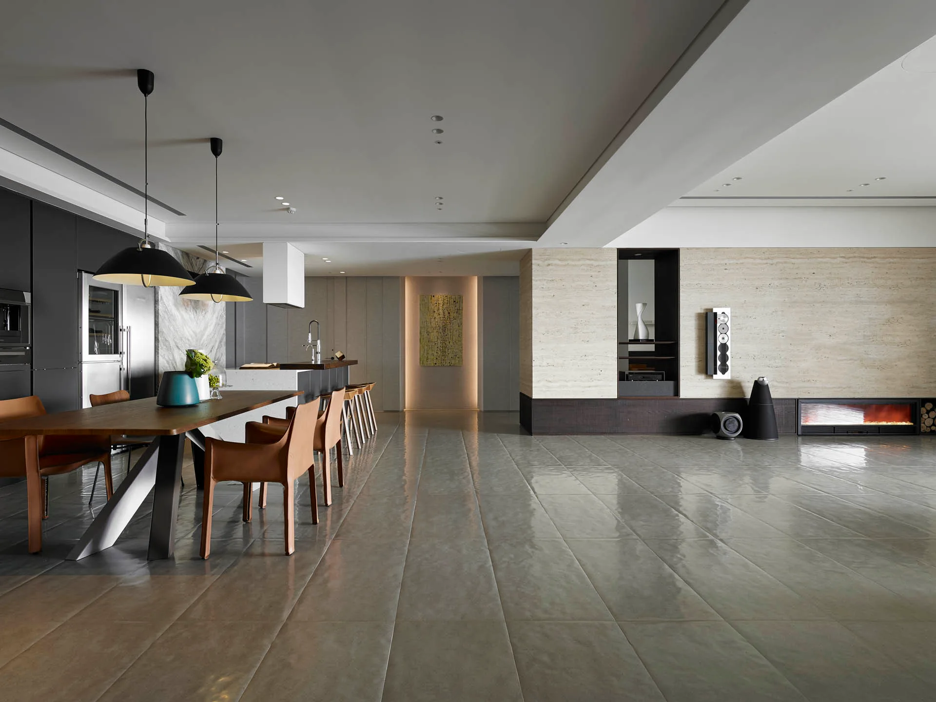 An open-concept kitchen and dining area features a large wooden dining table under black pendant lights next to a textured travertine feature wall.