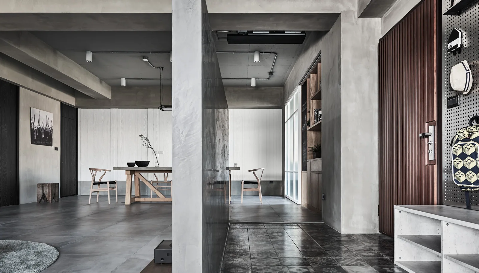 An industrial dining room featuring a central concrete partition wall, wooden dining table, wishbone chairs, and exposed concrete ceilings.