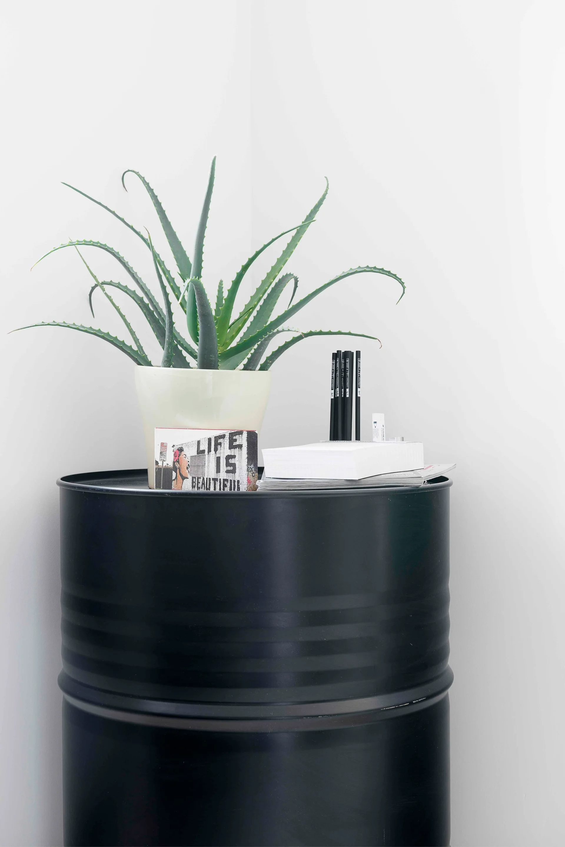 A repurposed black metal drum serves as a side table holding a potted aloe plant, a small card, and writing supplies against a white wall.