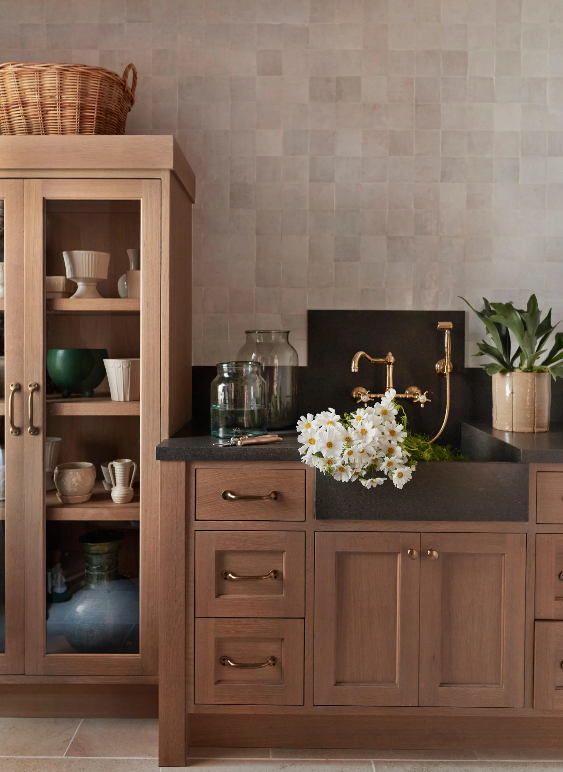 A light wood utility cabinet and dark stone sink with a brass wall-mounted faucet set against a square beige tile backsplash.