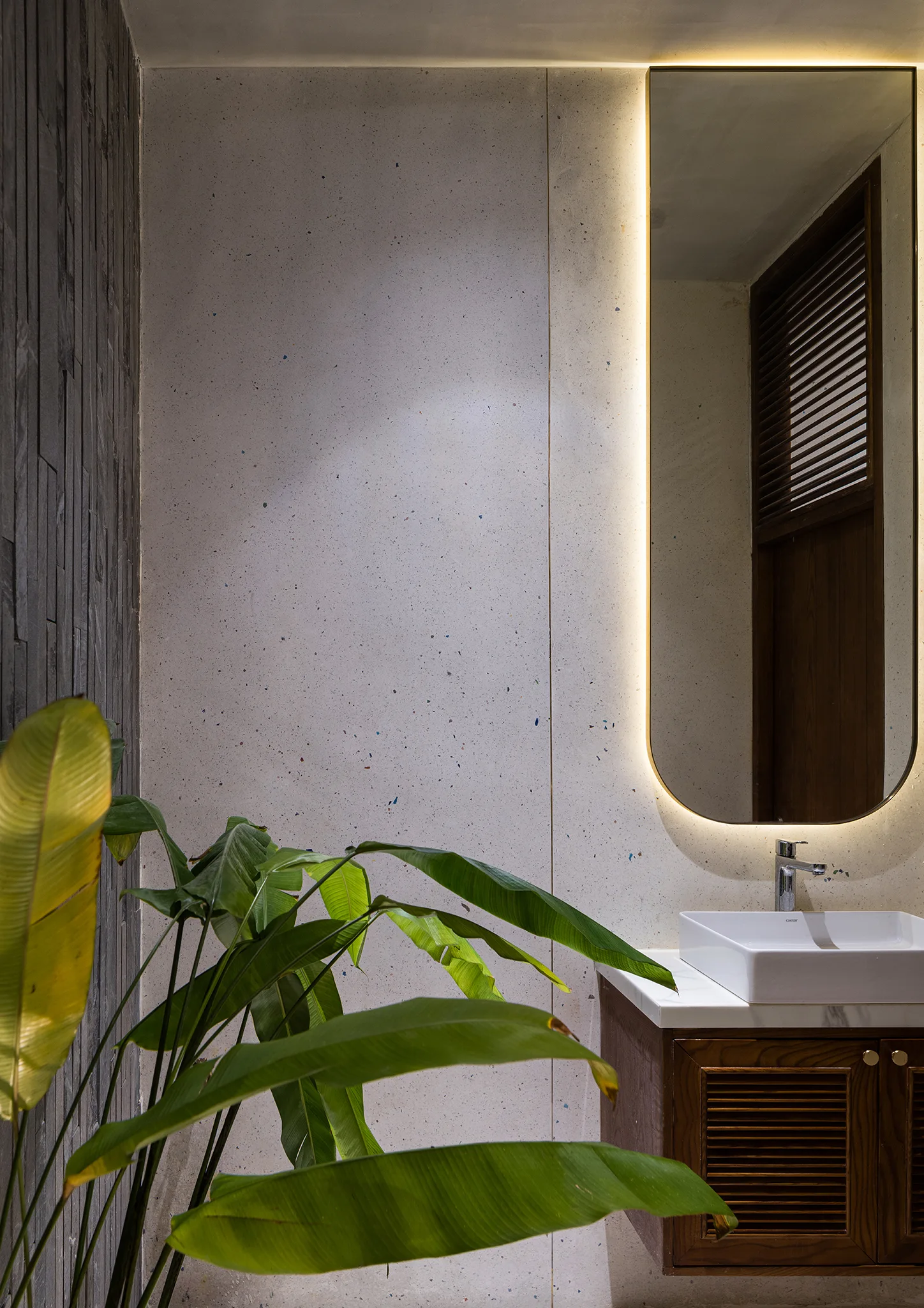 A modern bathroom vanity featuring a wooden louvered cabinet, a white ceramic sink basin, a tall backlit mirror, and a speckled wall.