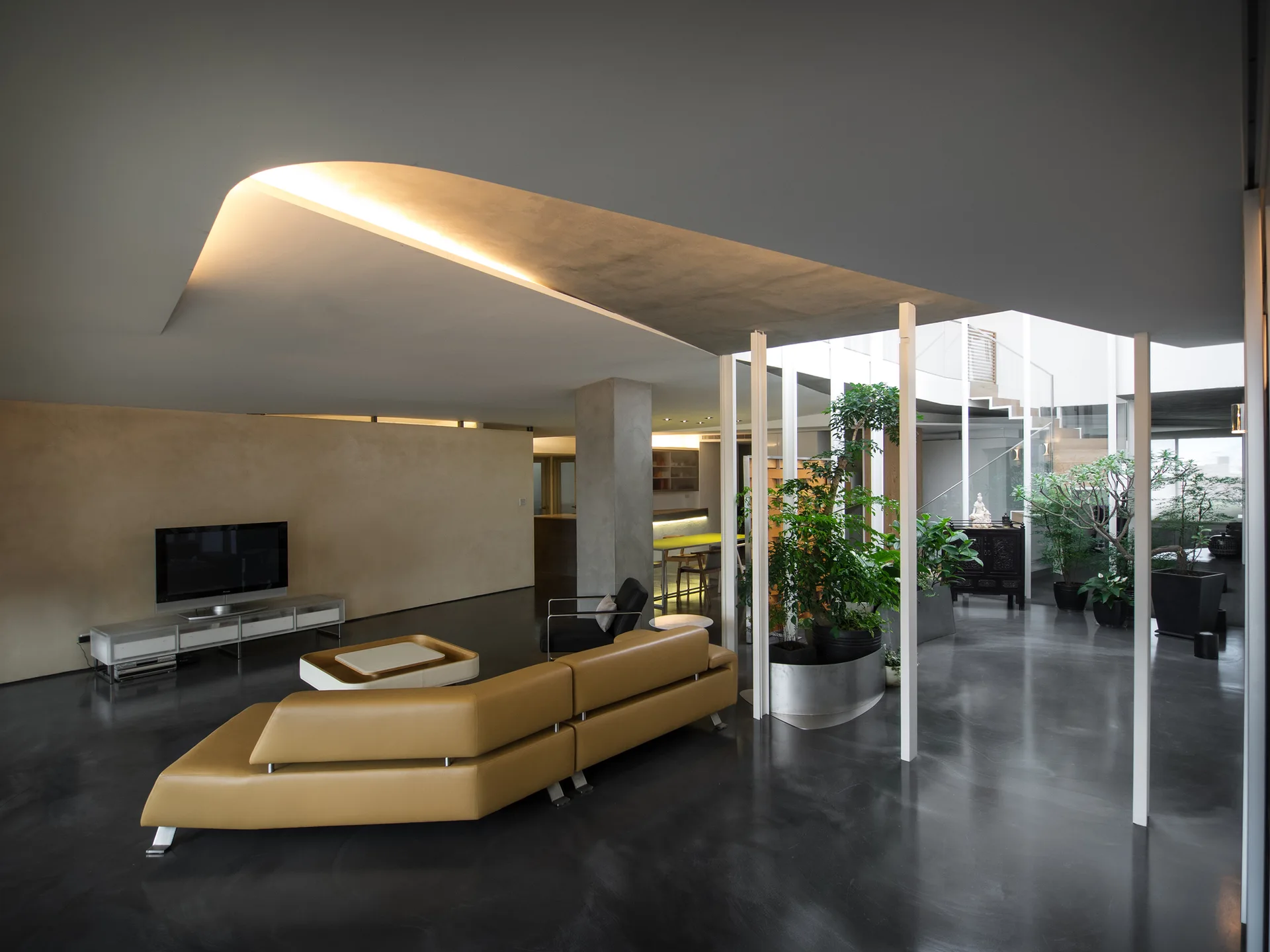 A minimalist living room with a tan leather sectional sofa, polished dark floors, and a coved ceiling with recessed lighting next to an indoor garden.