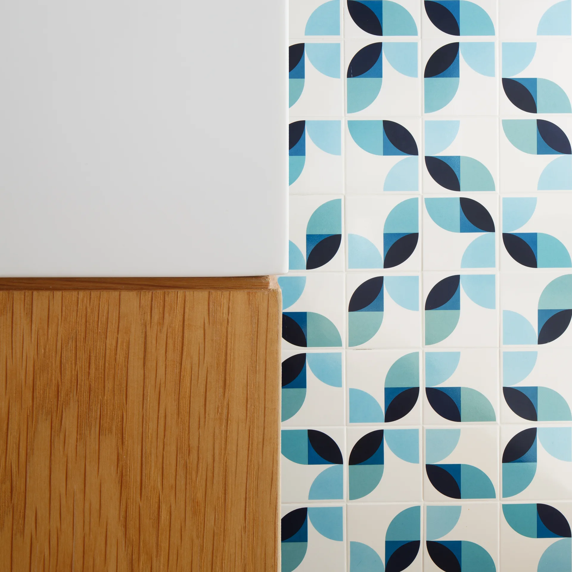 Glossy wall tiles with a blue and teal geometric petal pattern sit adjacent to a light wood cabinet panel and a plain white countertop.