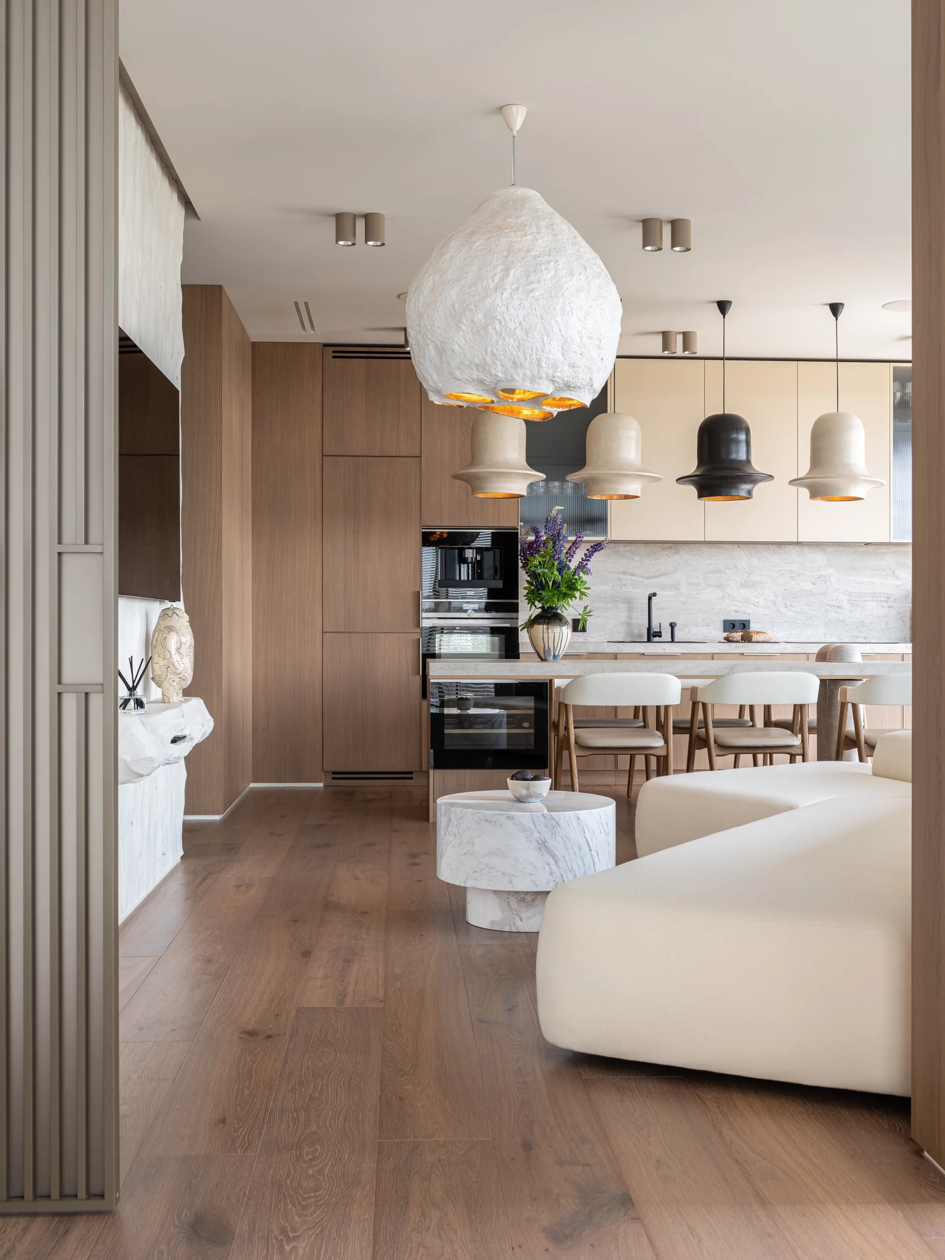 An open kitchen and living area featuring warm wood cabinetry, textured pendant lights over a stone island, and a curved cream sofa.