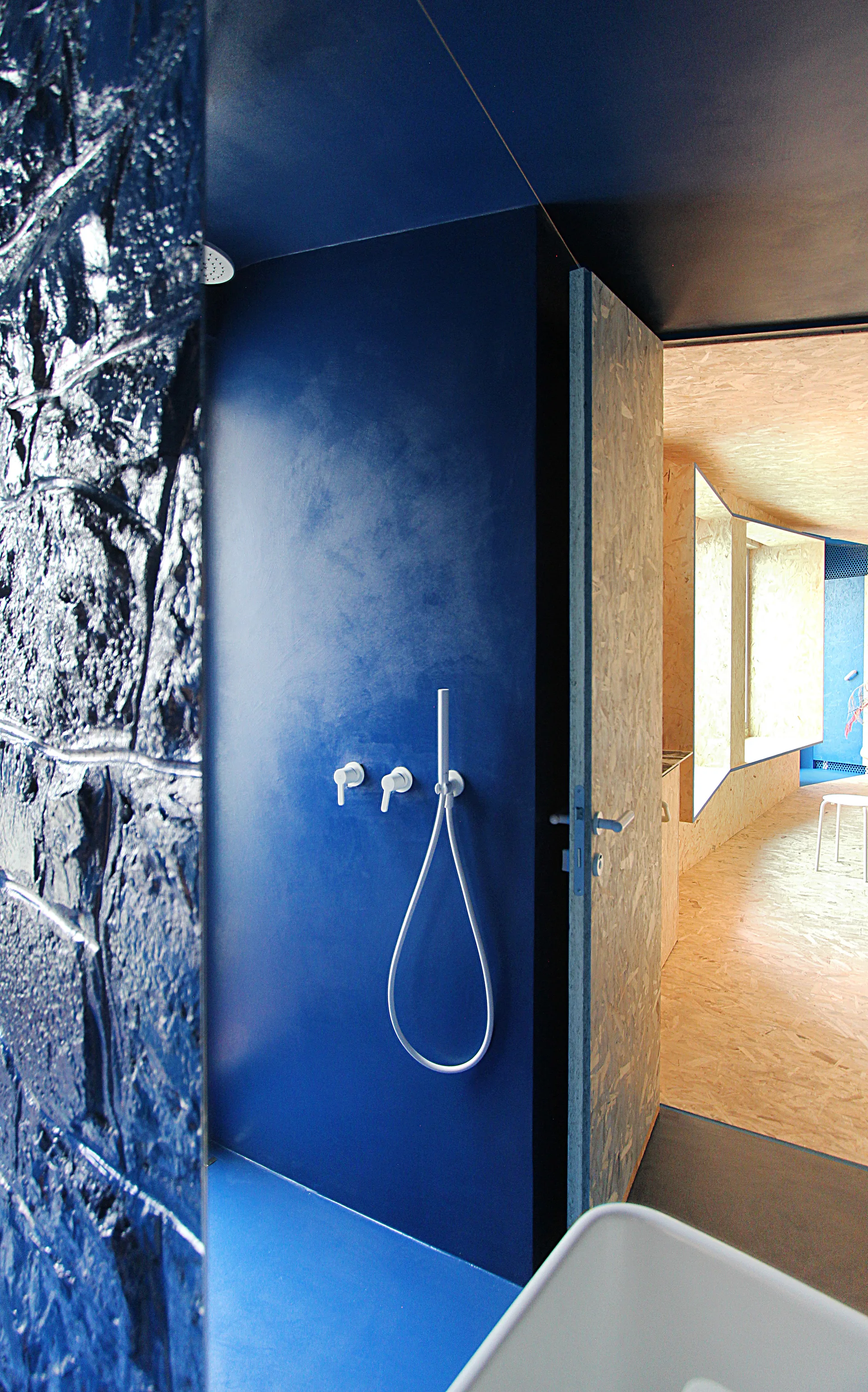 A modern bathroom features a deep blue matte shower with white fixtures, opening into an adjacent room walled with oriented strand board.