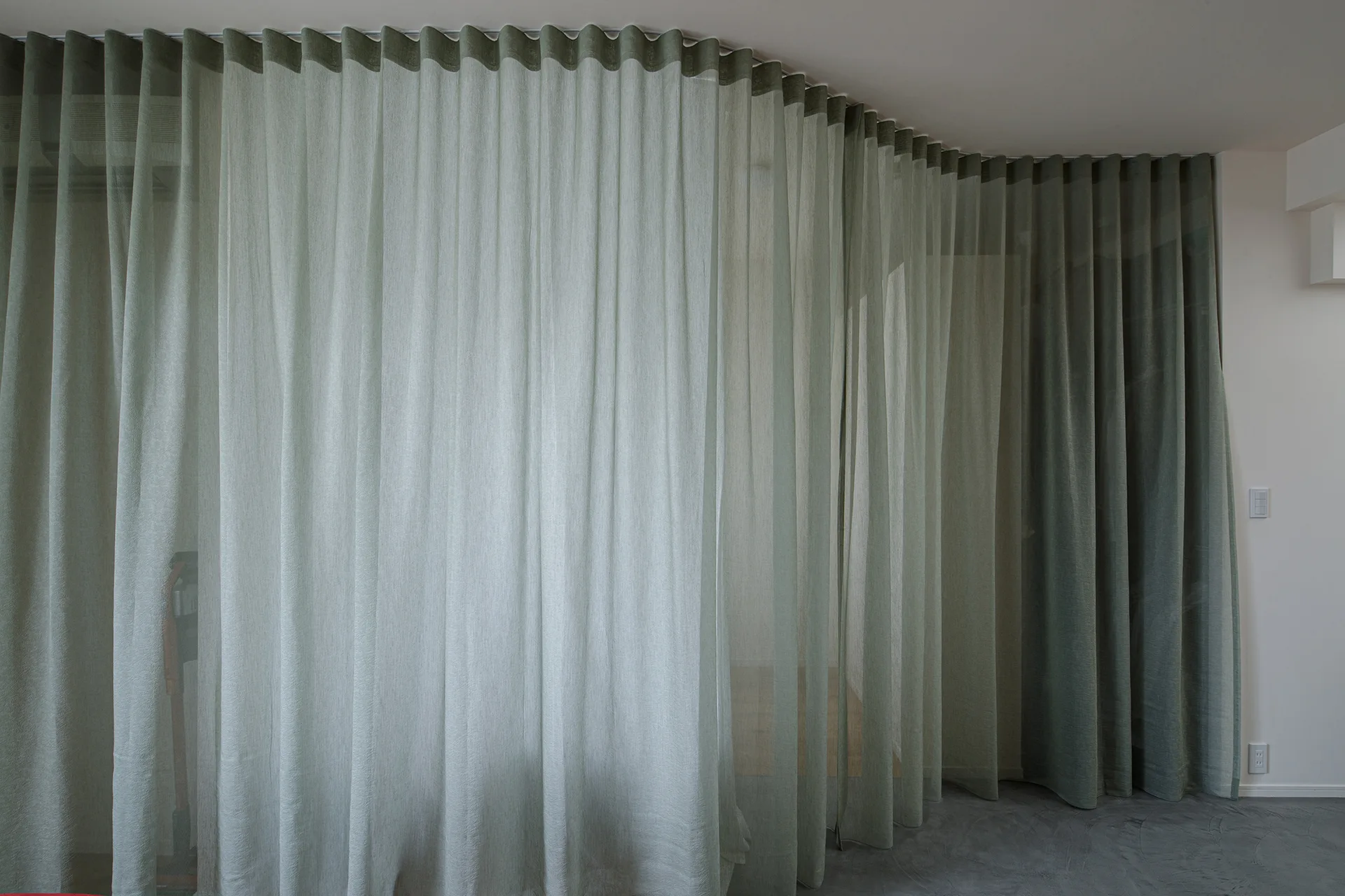 A floor-to-ceiling sage green sheer curtain hangs on a curved track, dividing a room with gray concrete floors and white walls.