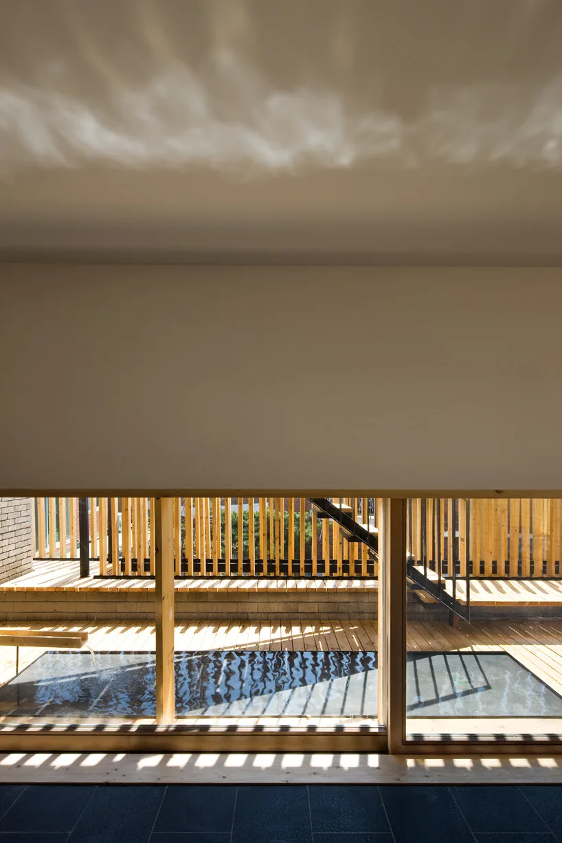 A view through a low window shows an outdoor wooden terrace with a shallow water feature and a vertical slatted privacy screen.