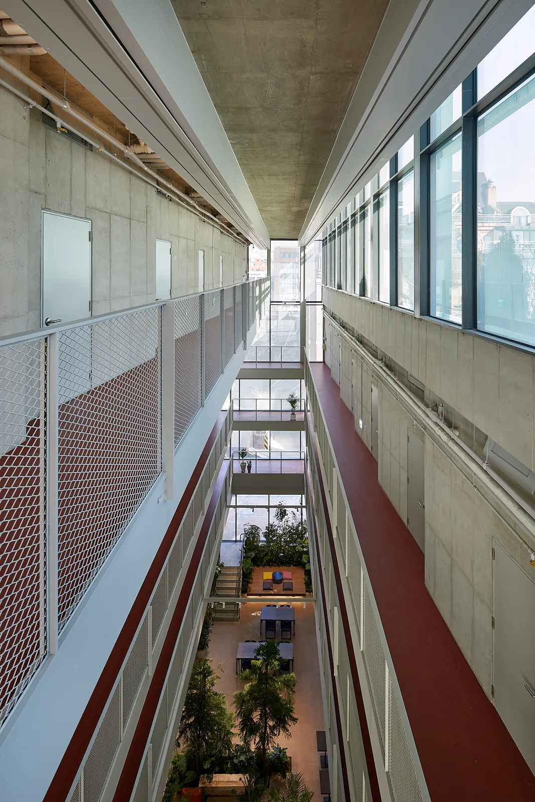 A multi-level interior atrium features exposed concrete walls, white metal mesh railings, red-floored walkways, and a central indoor courtyard.