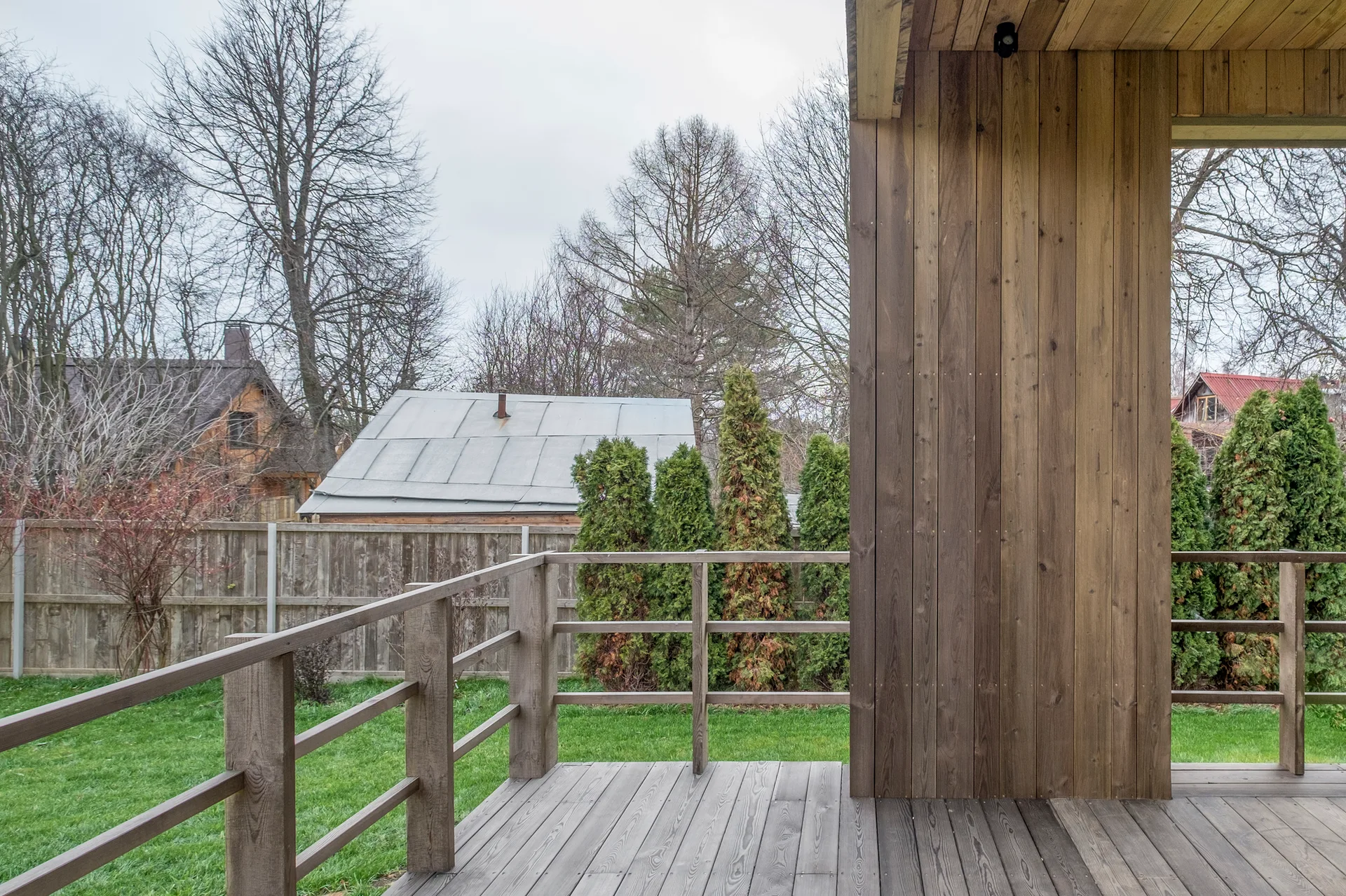 A stained wood deck with a simple wooden railing overlooks a grassy backyard with evergreen trees and a wooden fence.