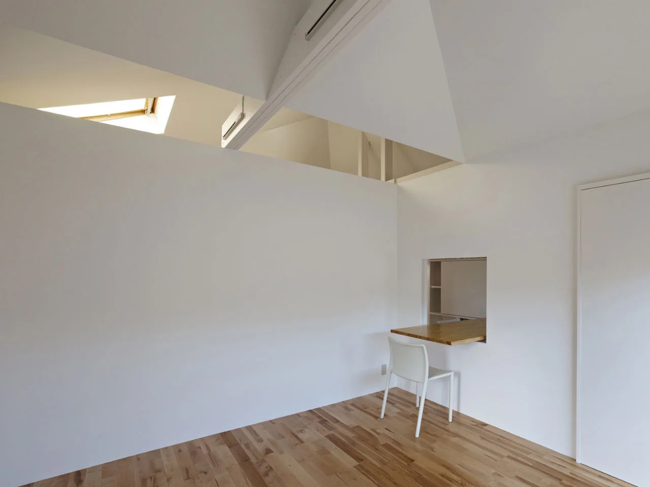 A minimalist white room with light wood floors features a wall-mounted wooden desk extending through a small wall cutout next to a white chair.