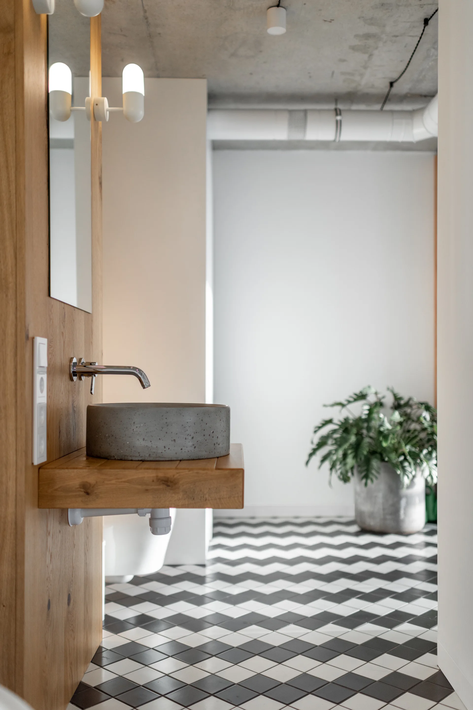 A concrete vessel sink sits on a floating wood vanity shelf in a bathroom with black and white chevron floor tiles and an exposed ceiling.