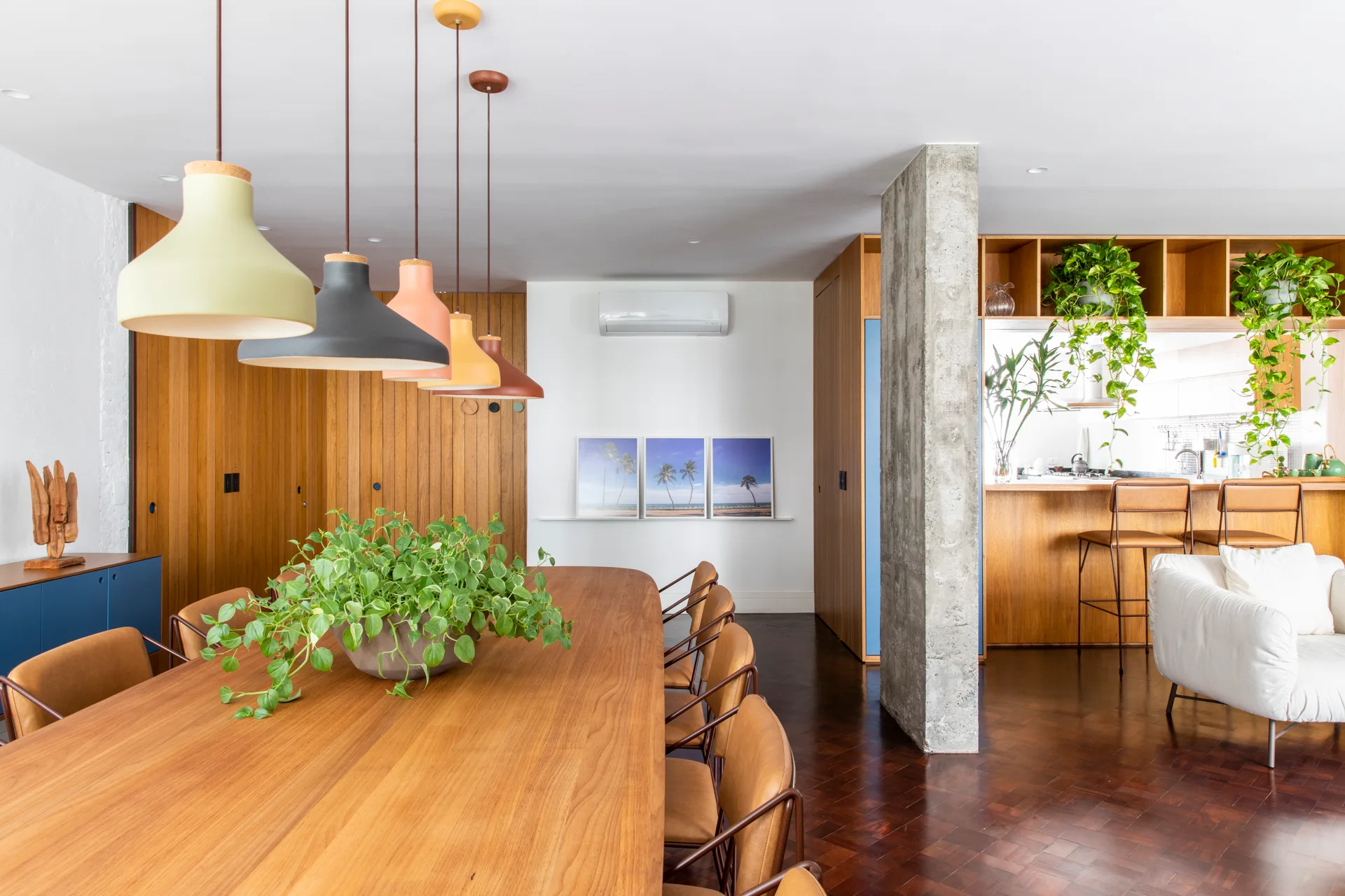 A modern dining area features a large wooden table with matching chairs, a row of multicolored pendant lights, and an exposed concrete column.