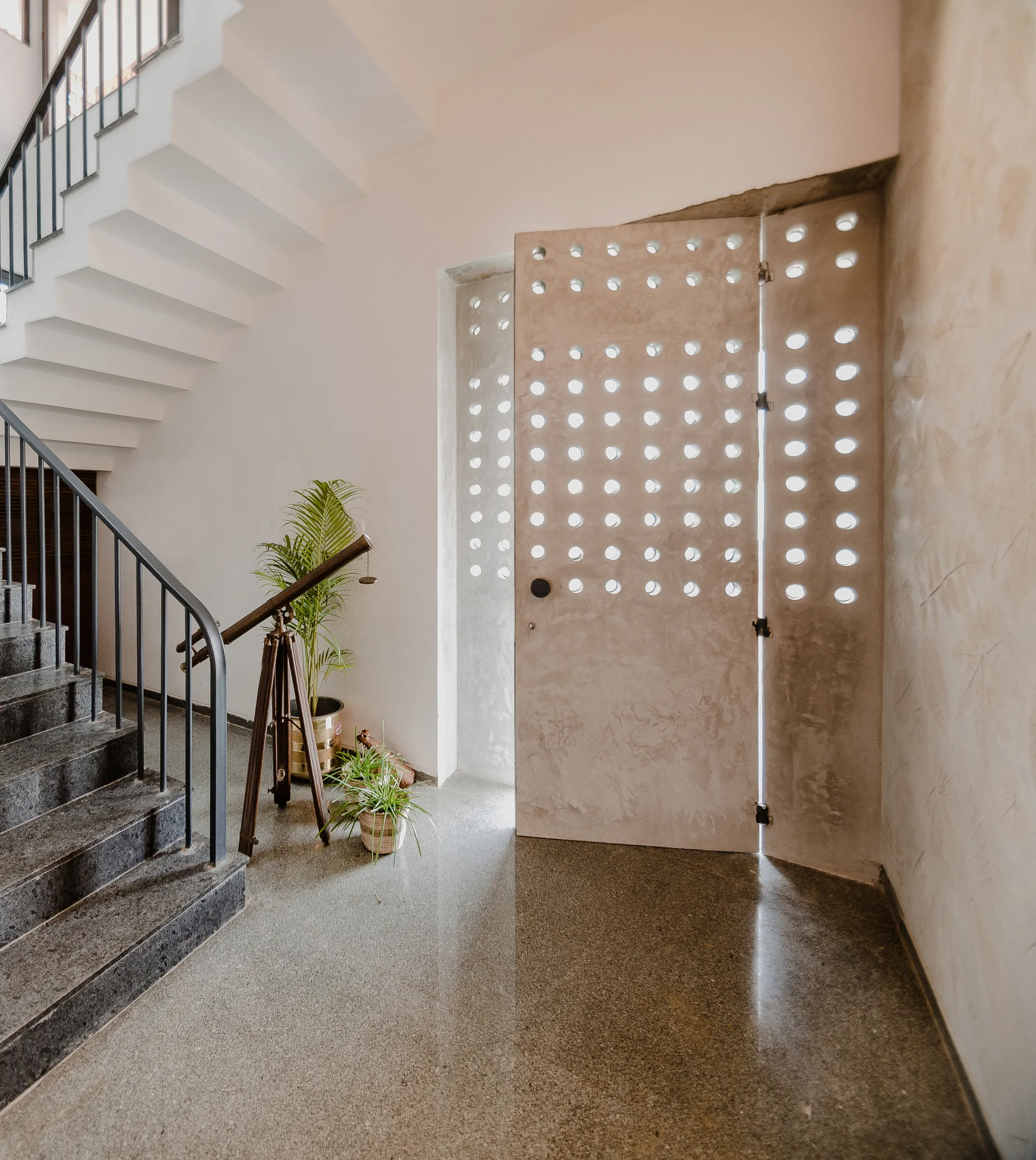 A modern minimalist entryway features a perforated concrete door with circular cutouts next to a dark stone staircase and polished flooring.