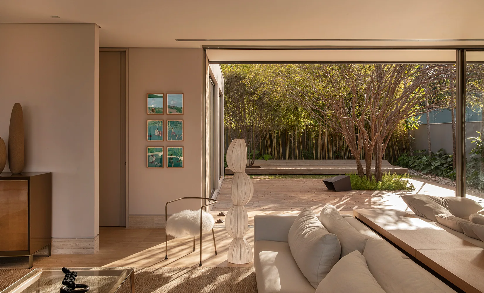 A modern neutral living room features a light fabric sofa, a sculptural floor lamp, and a large sliding door opening to a wooden terrace.