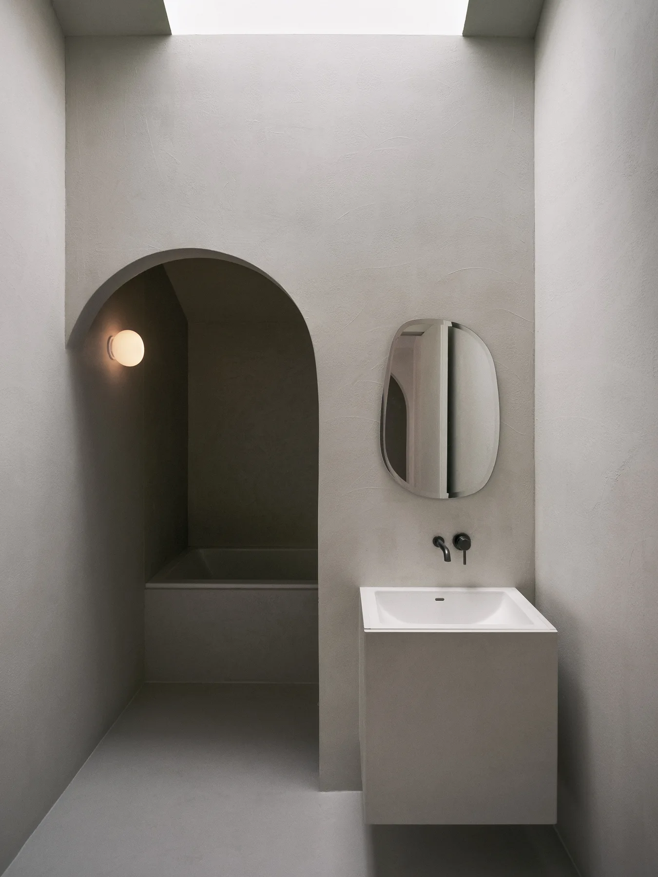 A minimalist bathroom features a freestanding greige sink vanity, an irregular wall mirror, and an arched entry to a recessed bathtub.