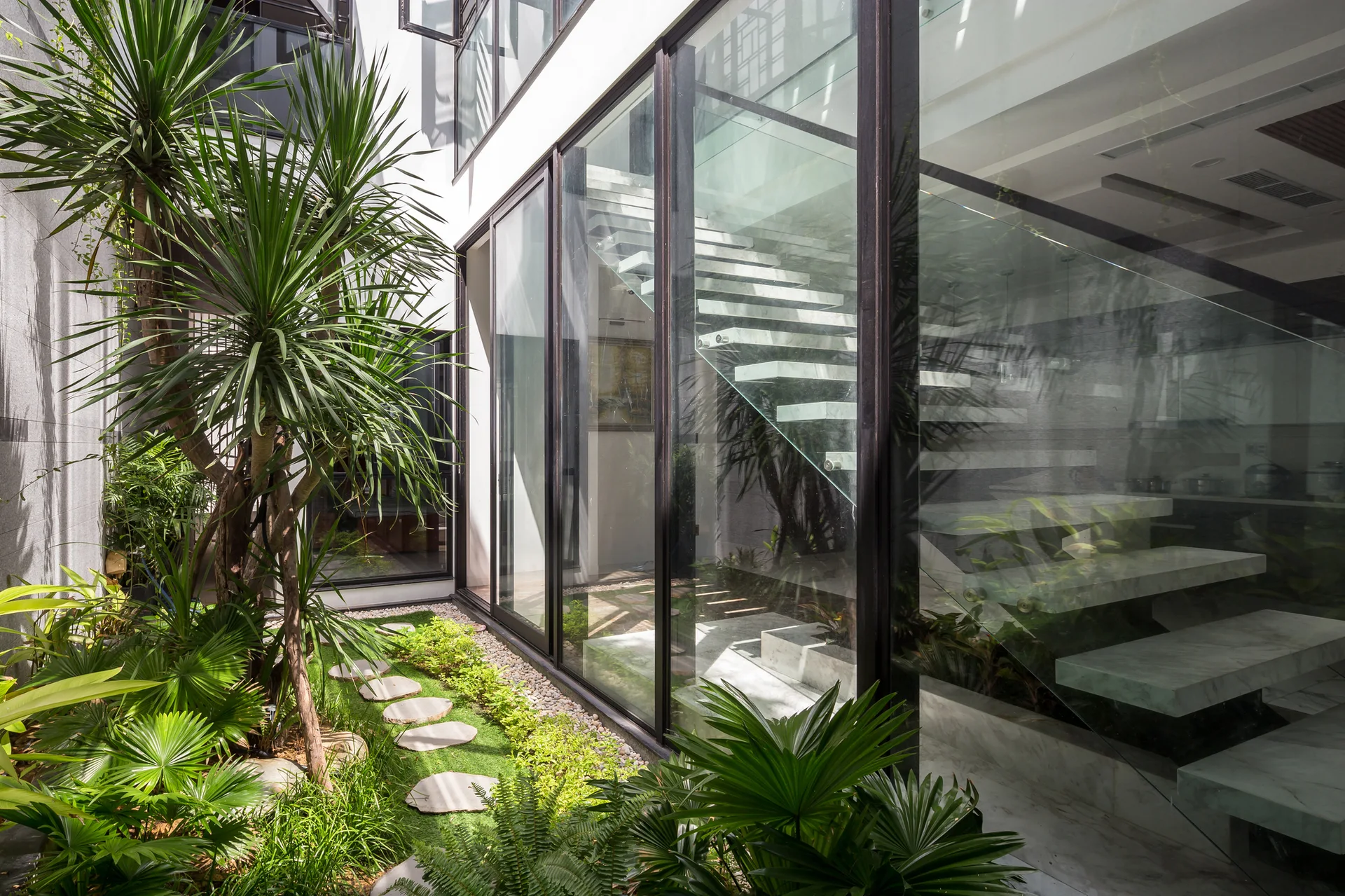 A modern floating stone staircase with glass railings is visible through a large glass wall next to a lush courtyard garden with stepping stones.