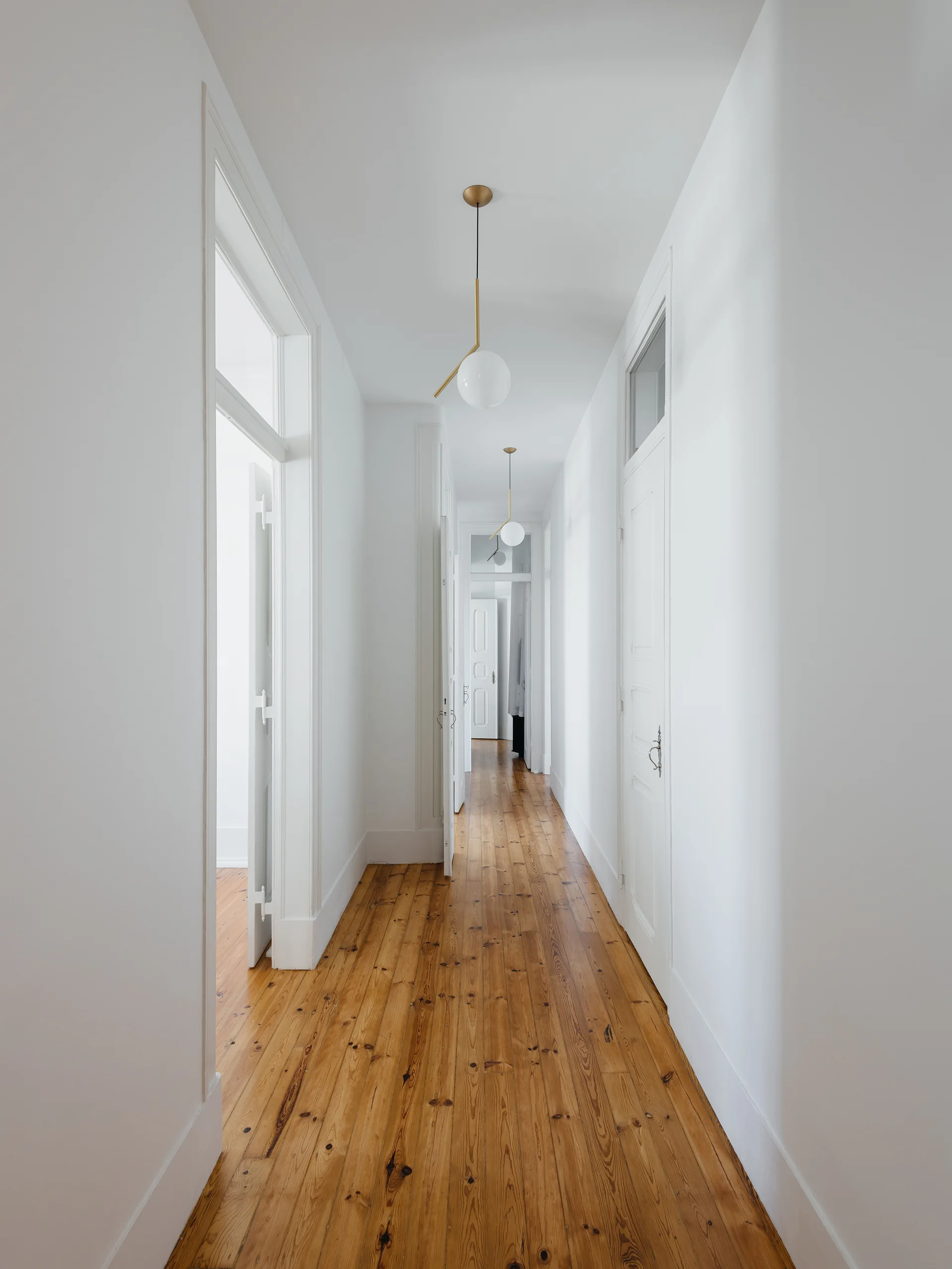 A long white hallway features natural wood plank flooring, multiple paneled doors with transom windows, and spherical brass pendant lights.