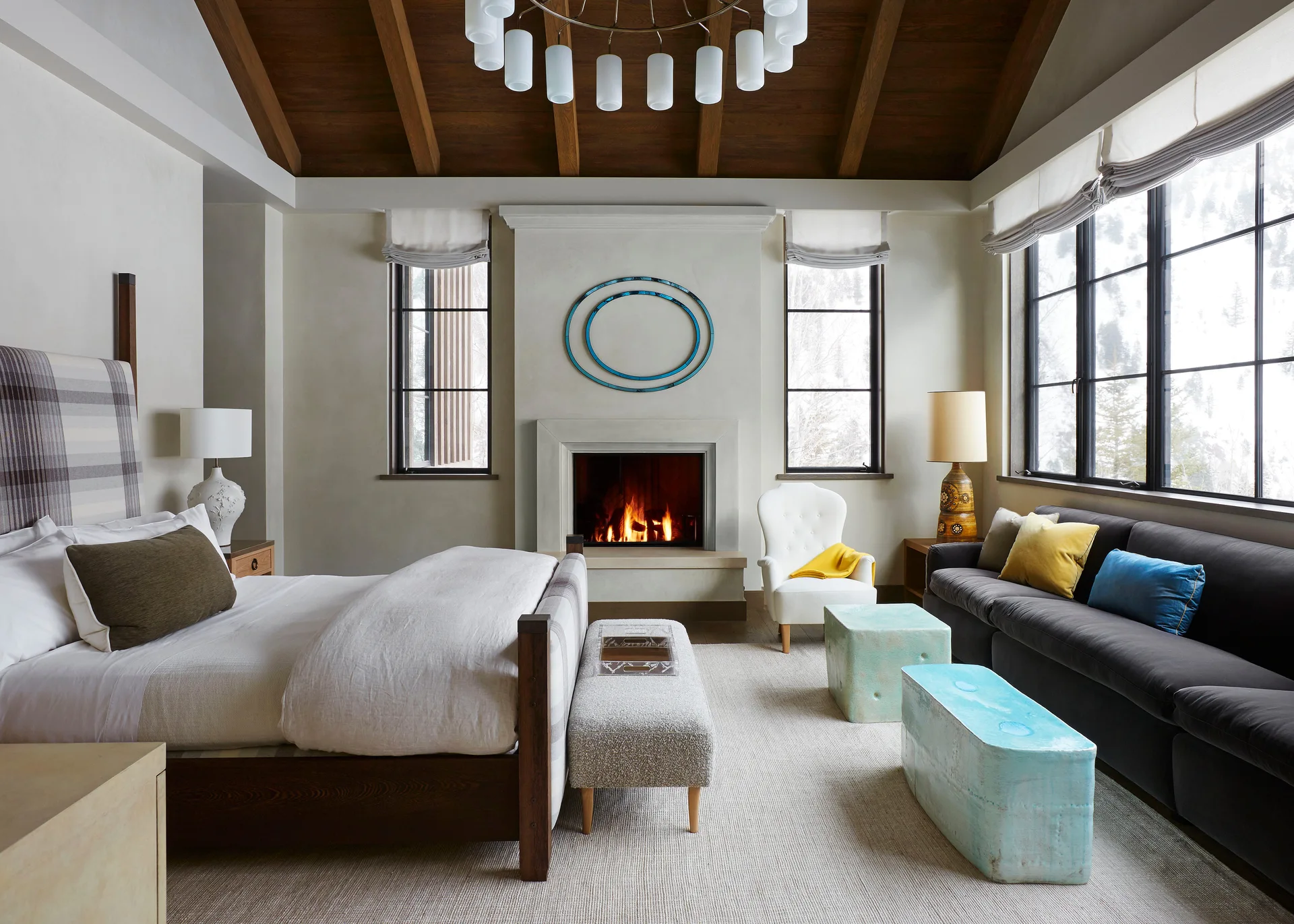 A spacious bedroom featuring a dark wood vaulted ceiling, a stone fireplace, a bed with a plaid headboard, and a sitting area with a dark sofa.