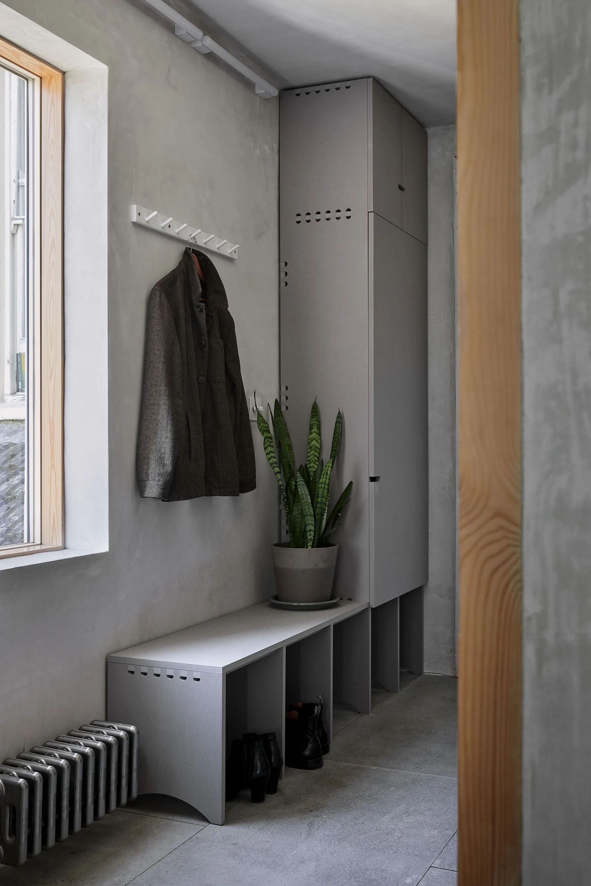 A minimalist gray mudroom featuring a built-in storage locker, a low bench with shoes underneath, a coat on a wall rack, and a potted plant.