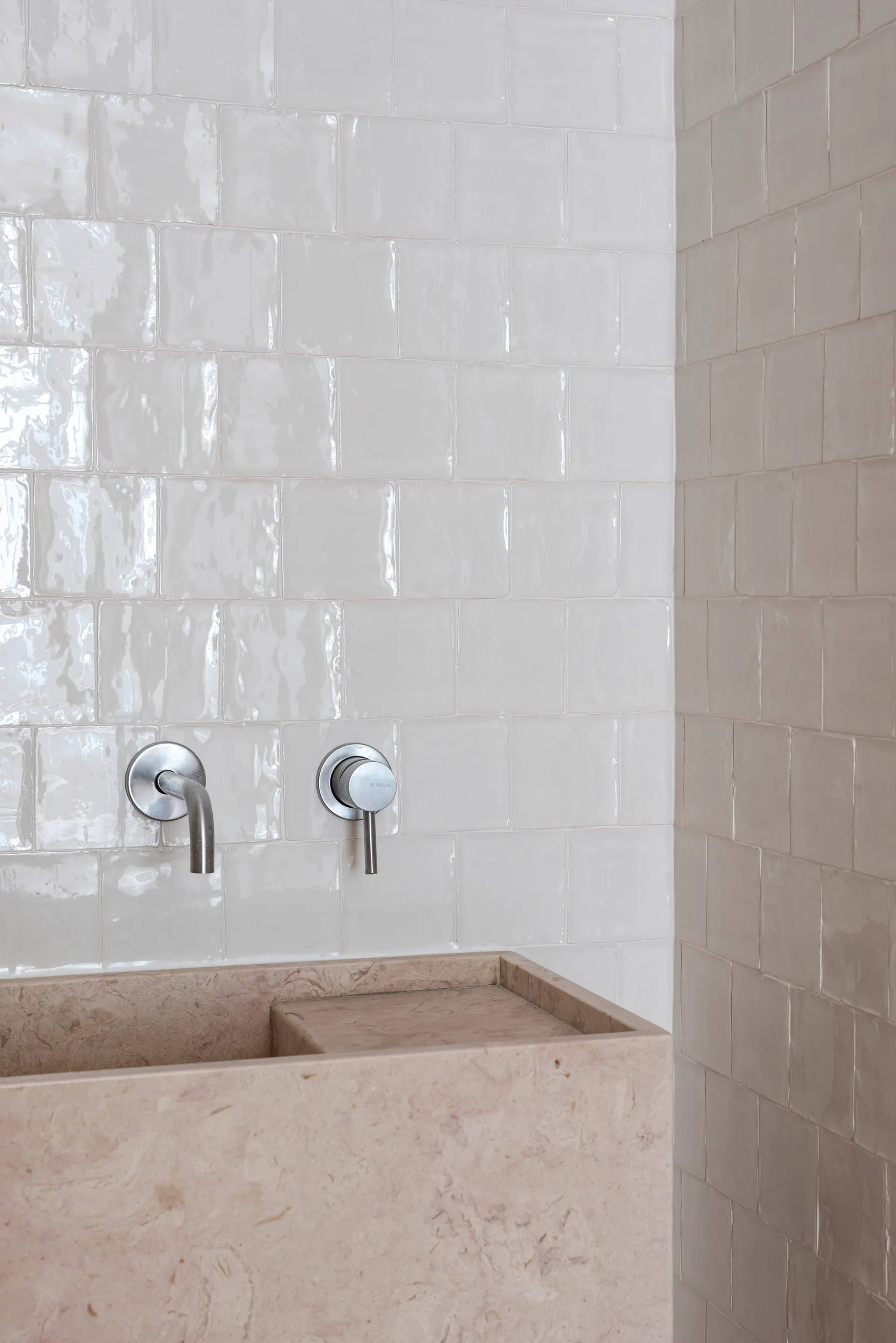 A textured pinkish-beige stone sink basin with a wall-mounted metal faucet sits against a backdrop of glossy square tiles.