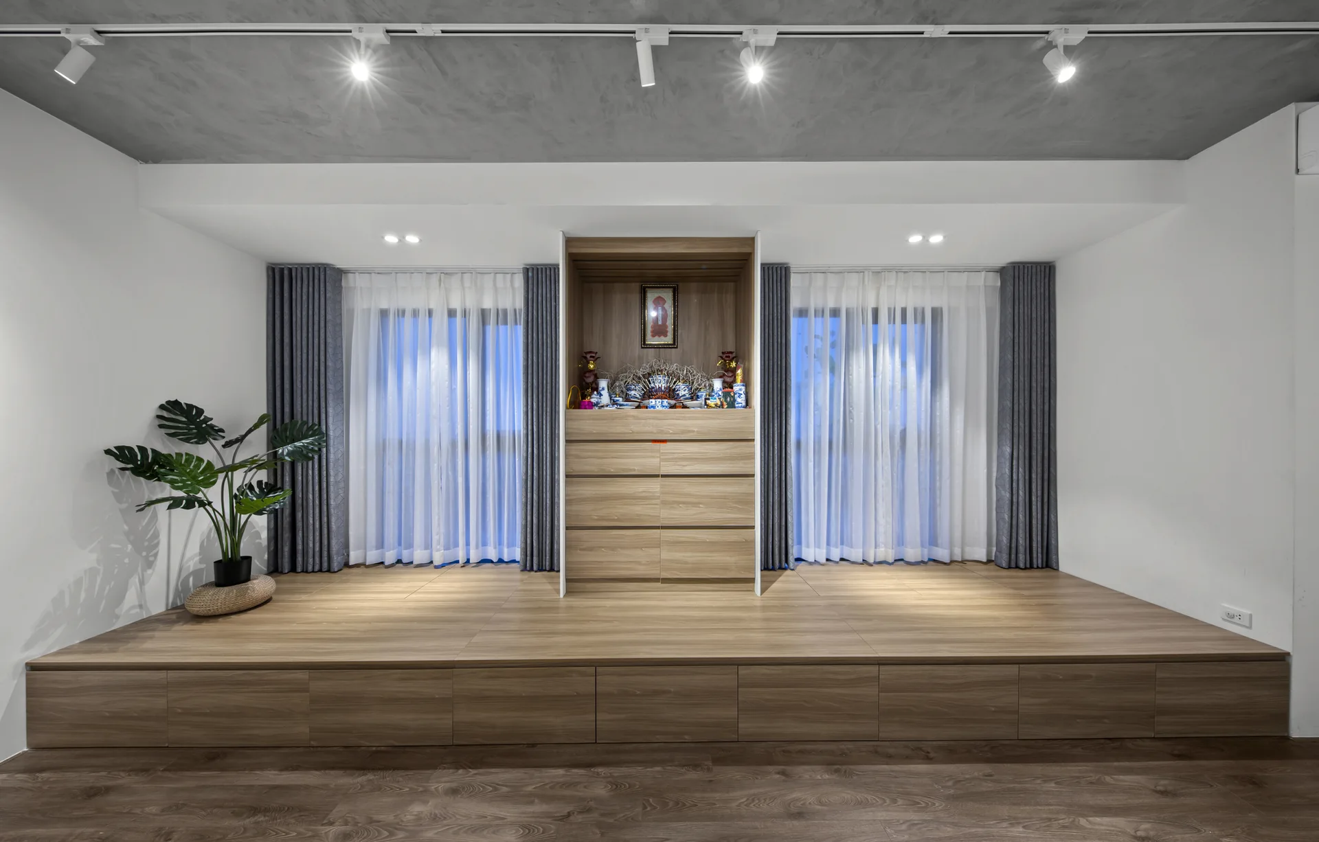 A modern room featuring a raised wood storage platform with built-in drawers, a central wooden altar cabinet, and draped windows.