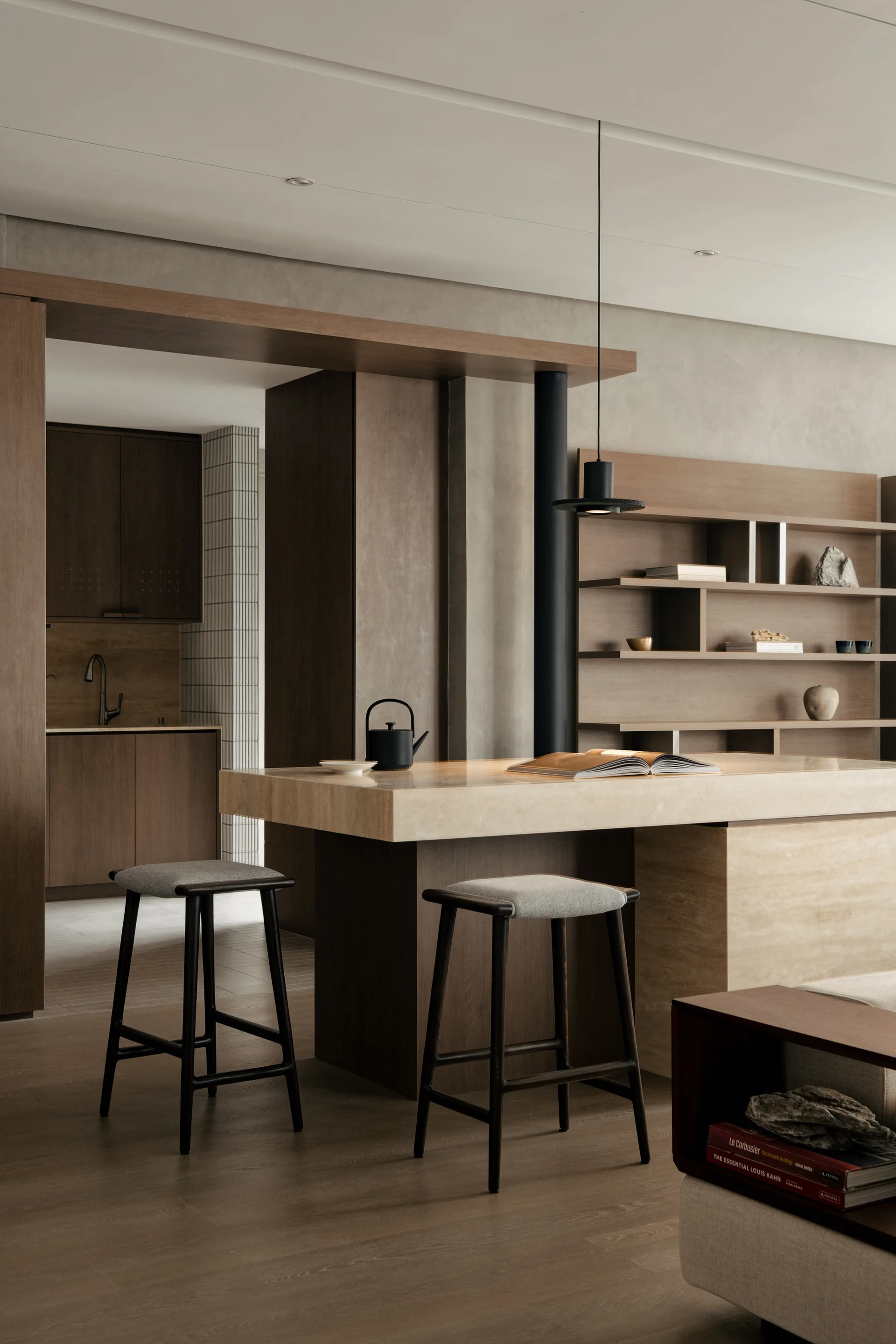 A minimalist kitchen space featuring a thick travertine stone island, two dark wood bar stools, and a large built-in wooden shelving unit.