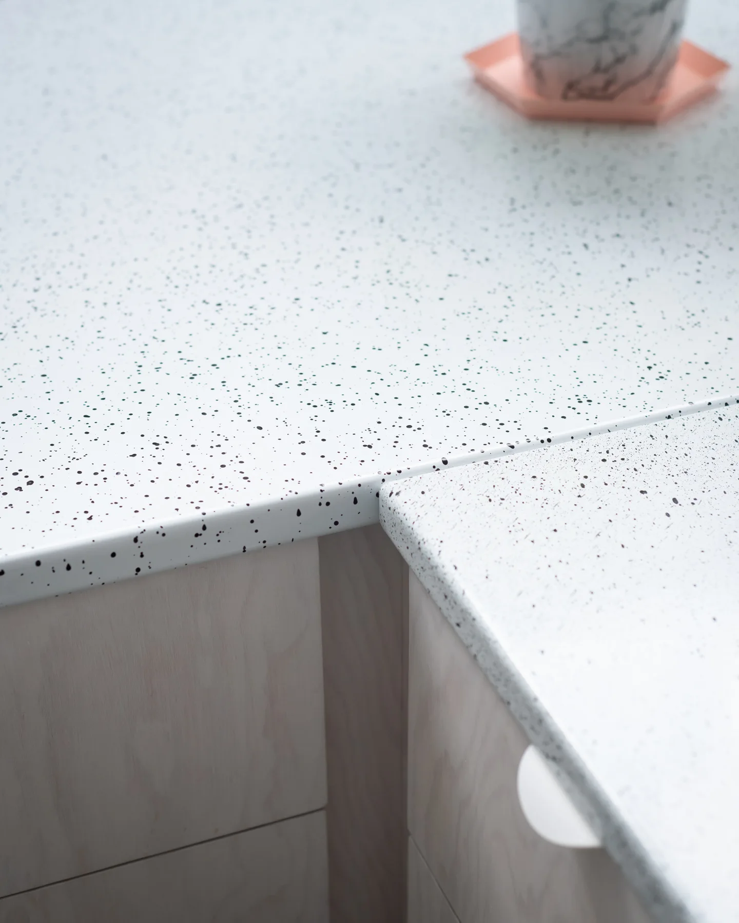 A close-up of a matte white countertop with black speckles meeting at a corner joint above pale wood kitchen cabinets.