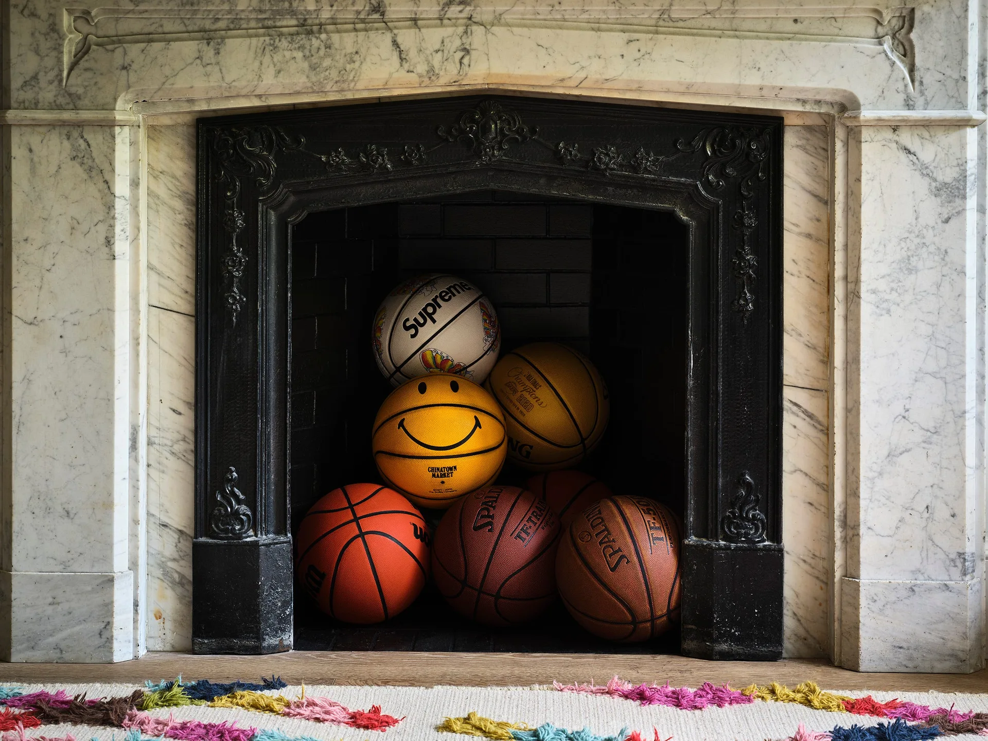 A pile of colorful, branded basketballs is stacked inside the dark hearth of an ornate white marble fireplace.