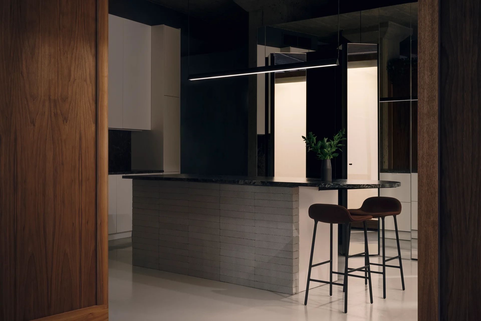 A minimalist kitchen with a light grey brick-textured island, dark stone countertop, two bar stools, and a sleek linear pendant light.